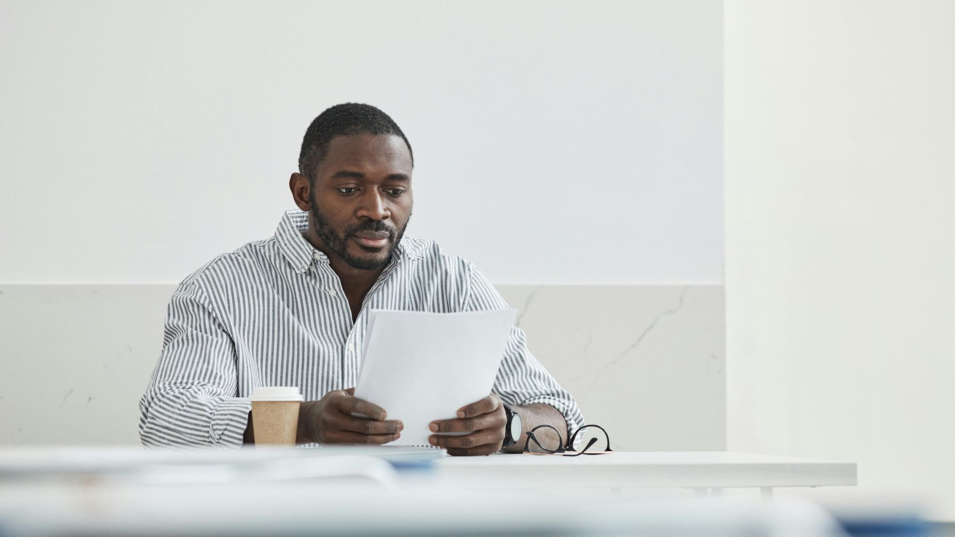 African American man sitting indoors, reading papers with a coffee cup nearby.
