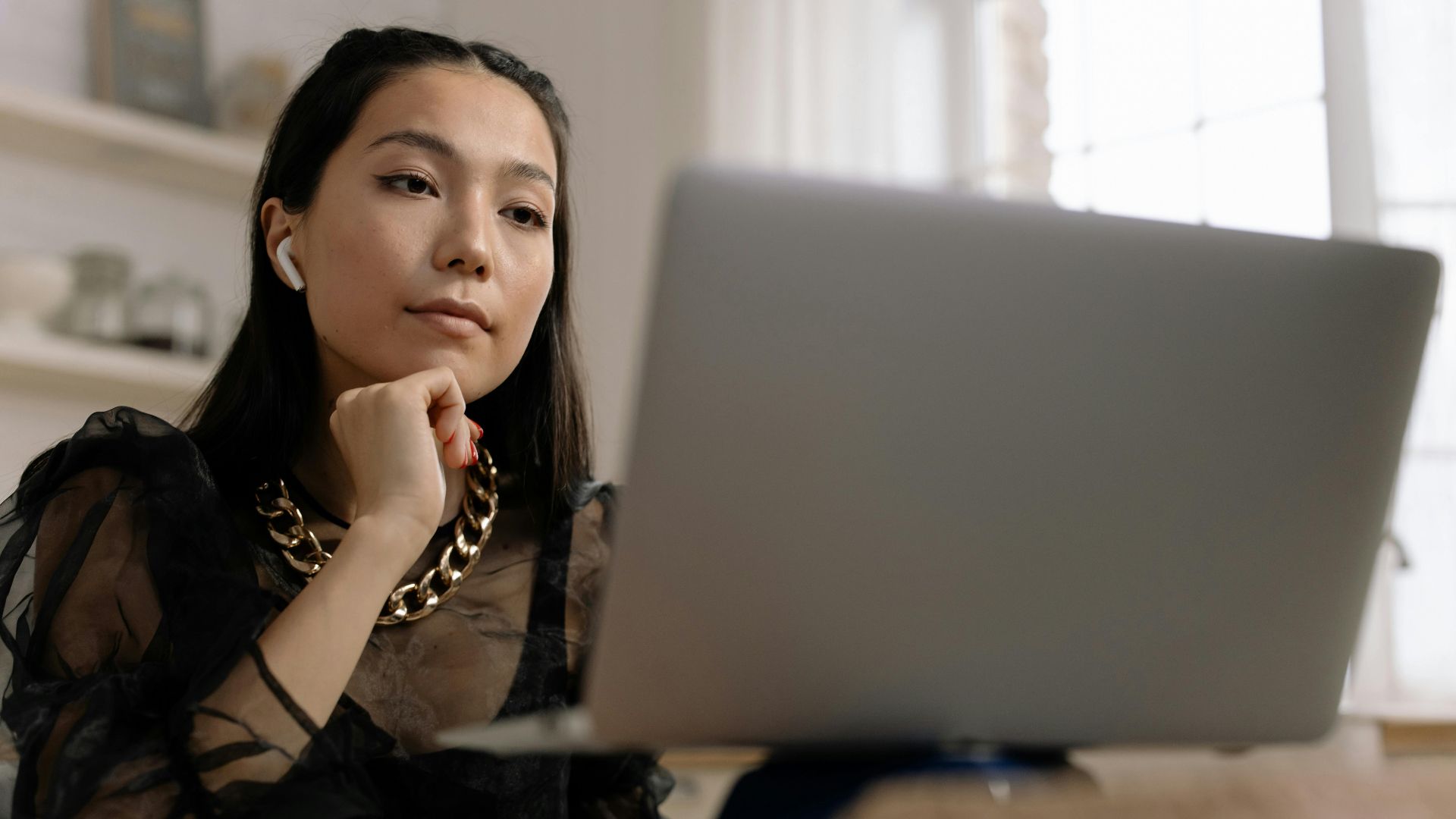 Young woman with wireless earphones working on a laptop indoors, displaying concentration and style.