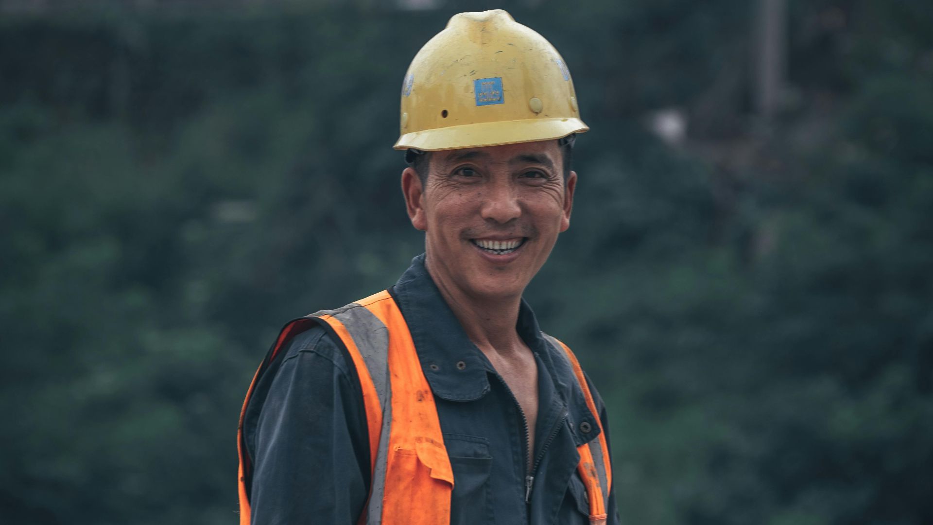 A smiling construction worker wearing safety gear stands outdoors in a work environment.
