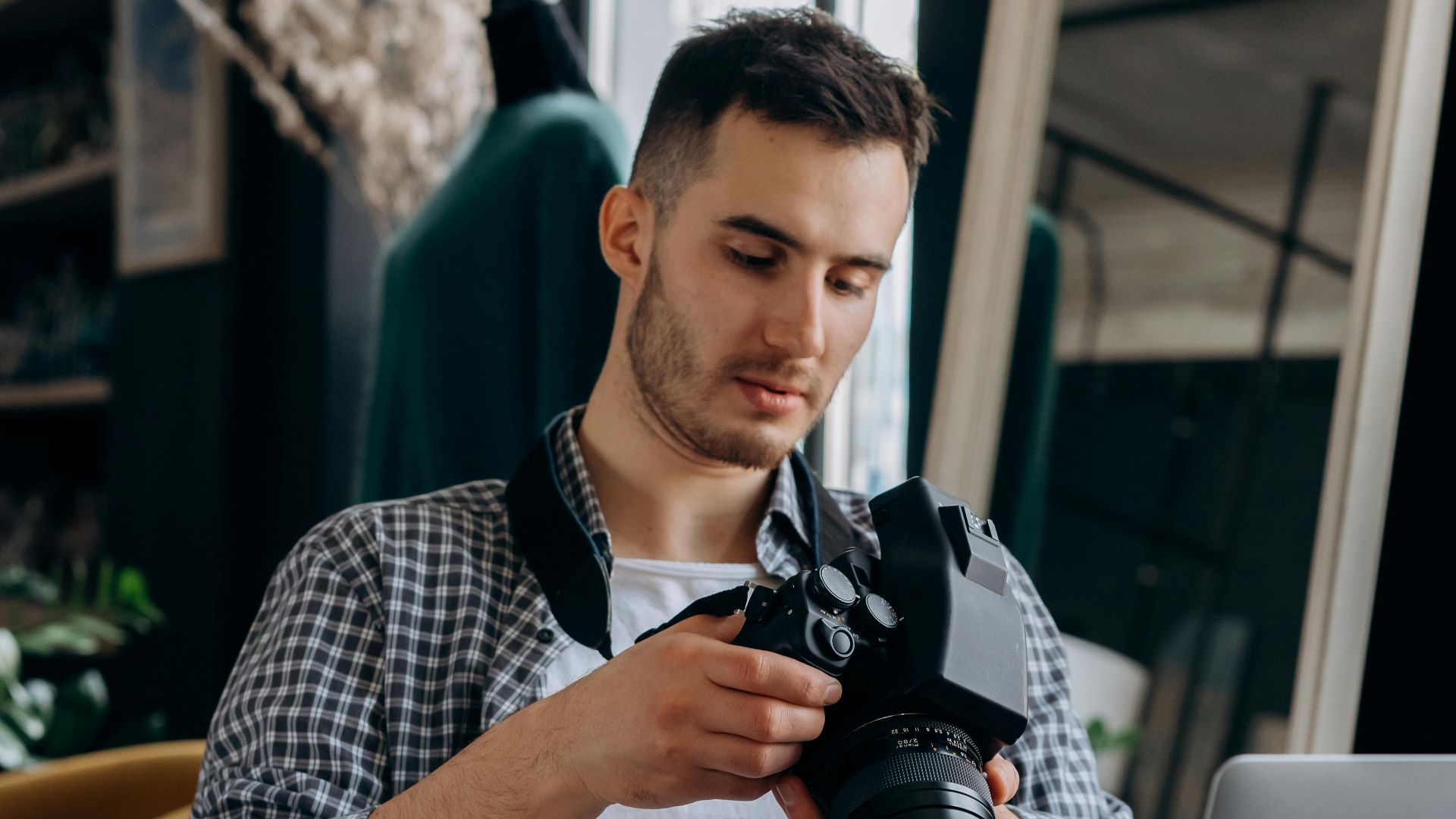 Young caucasian man checking DSLR camera settings while sitting indoors, near a laptop.