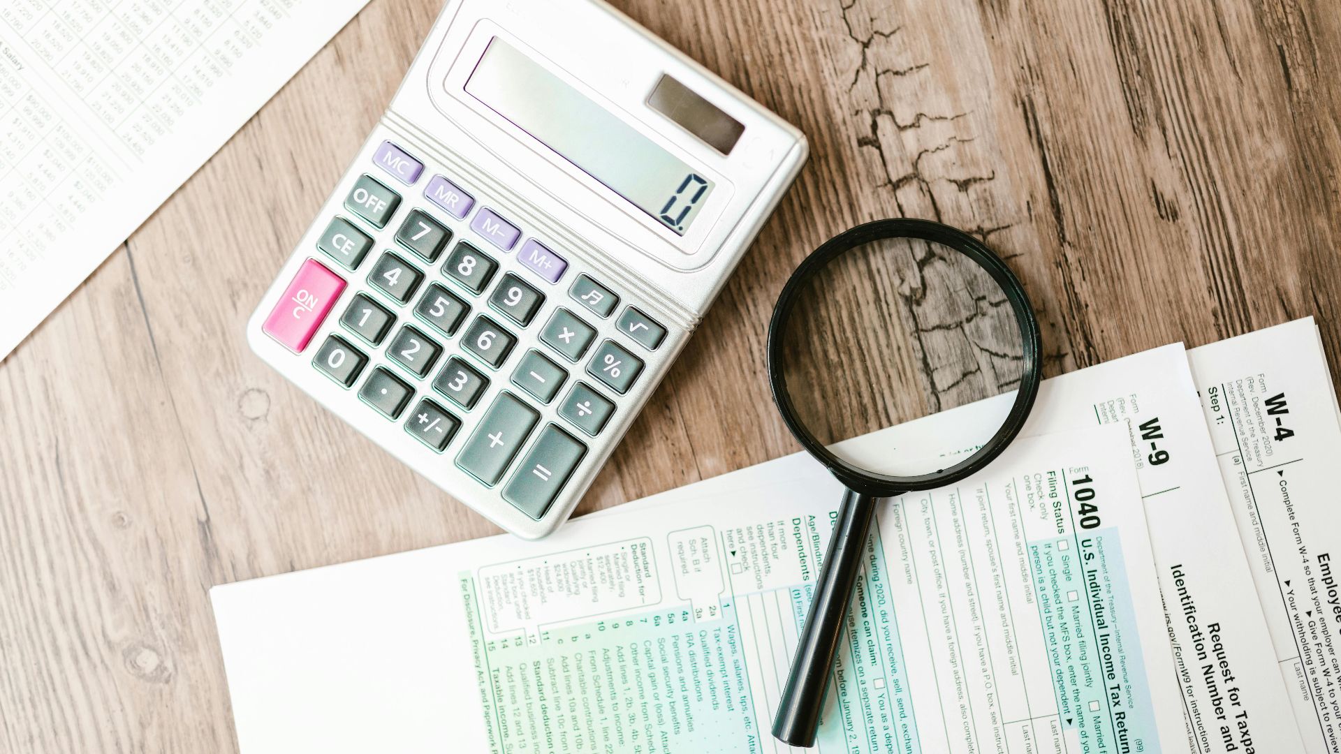 Tax forms laid out with a calculator and magnifying glass on a wooden surface, perfect for finance themes.