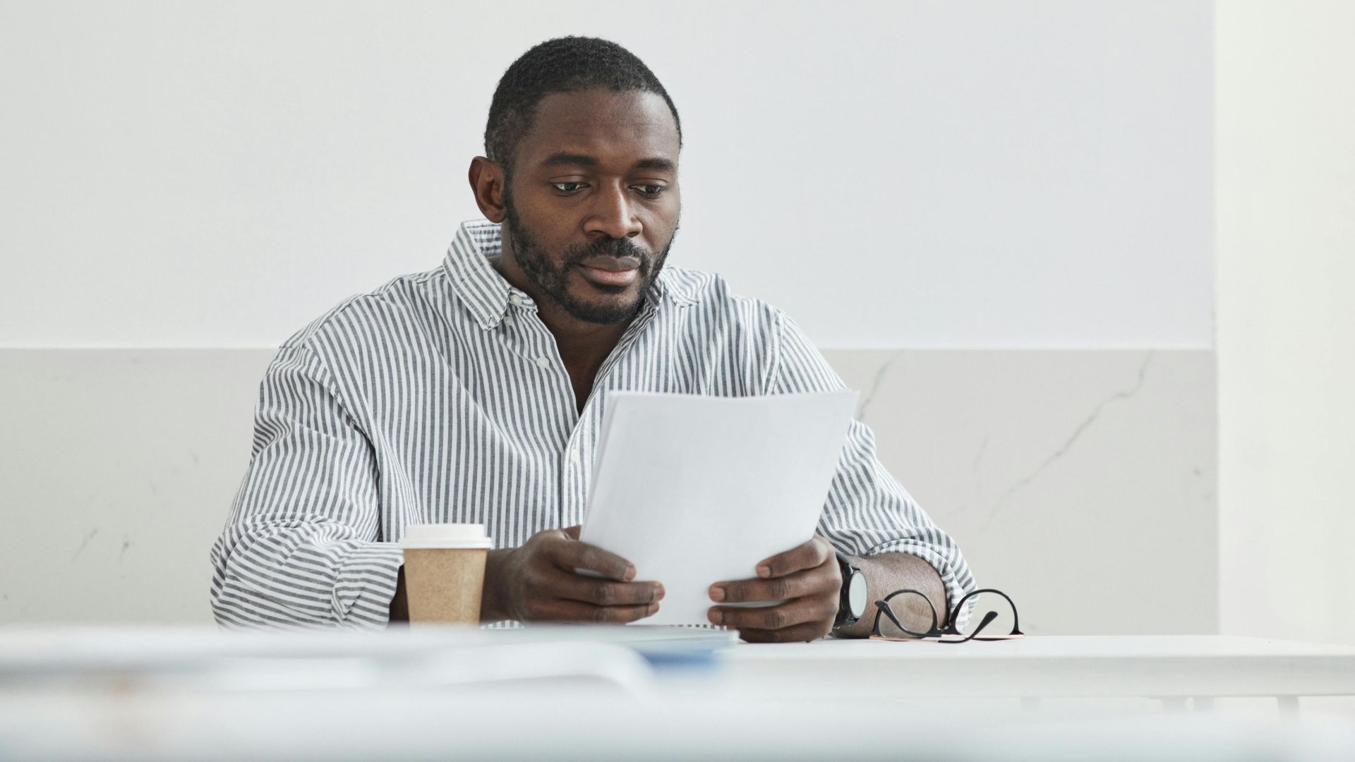 African American man sitting indoors, reading papers with a coffee cup nearby.
