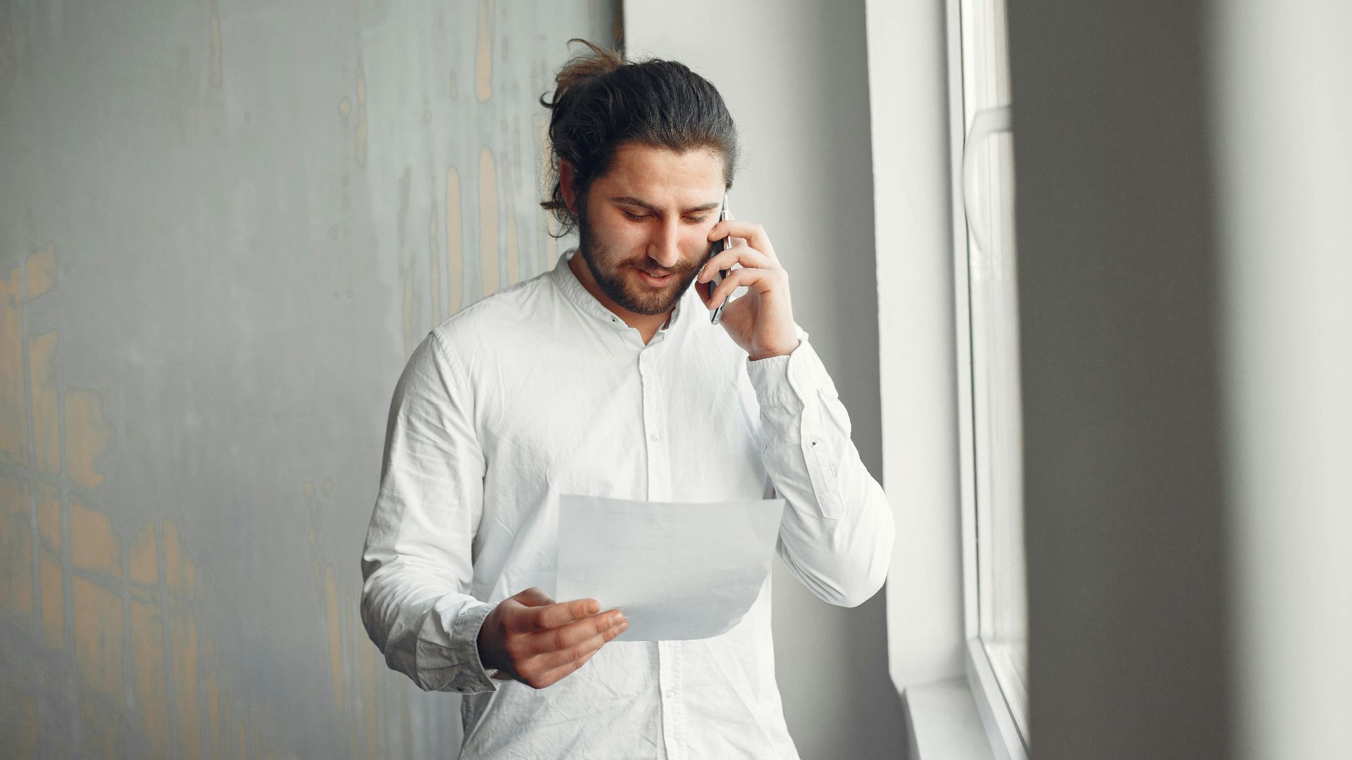 A young man in casual attire talks on the phone while holding a document indoors.