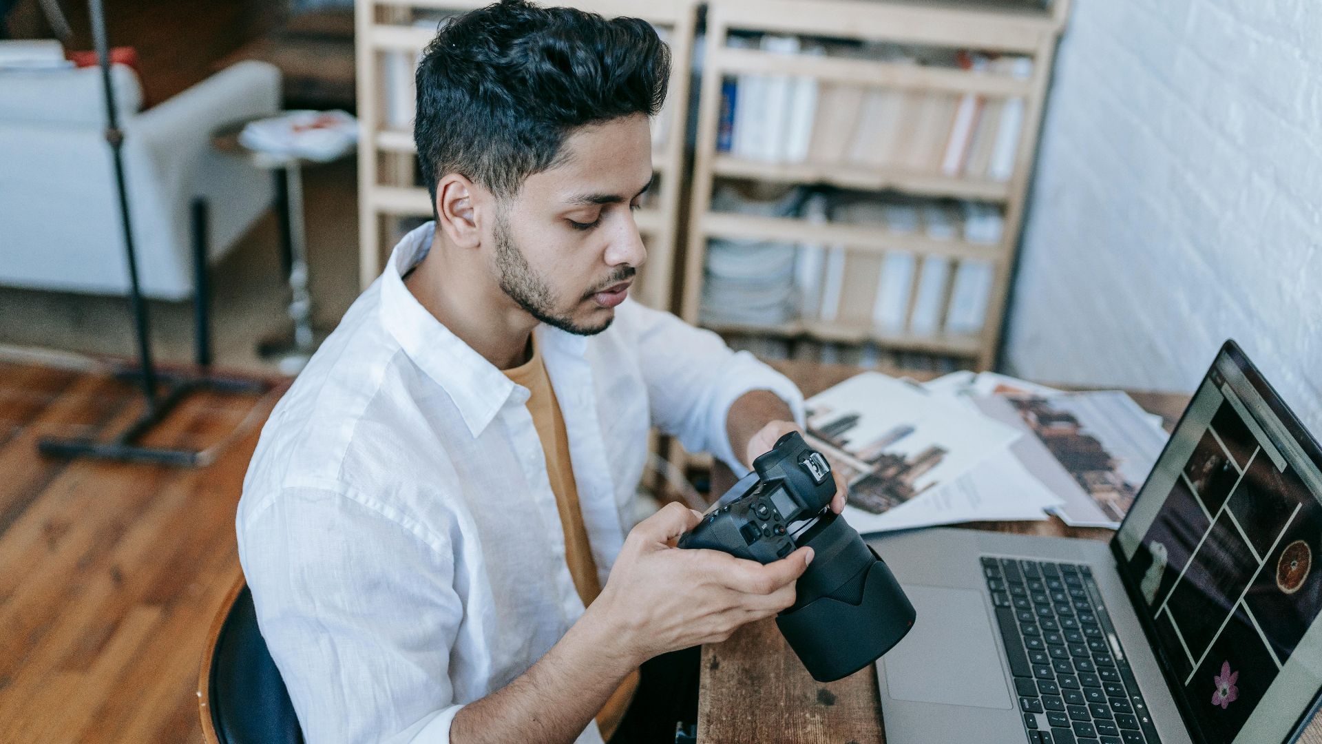 Side view of Indian male photographer in casual clothes watching photos on photo camera while sitting at table and browsing laptop at home in daytime