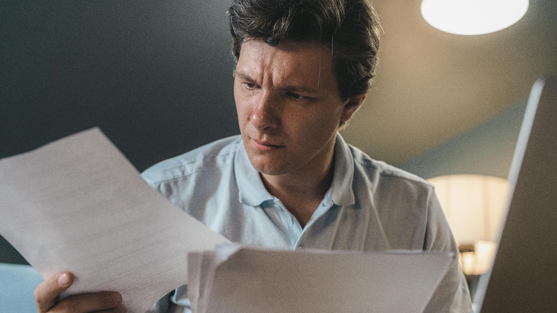 A man intently examines papers, seated indoors under warm lighting, focusing on his work.