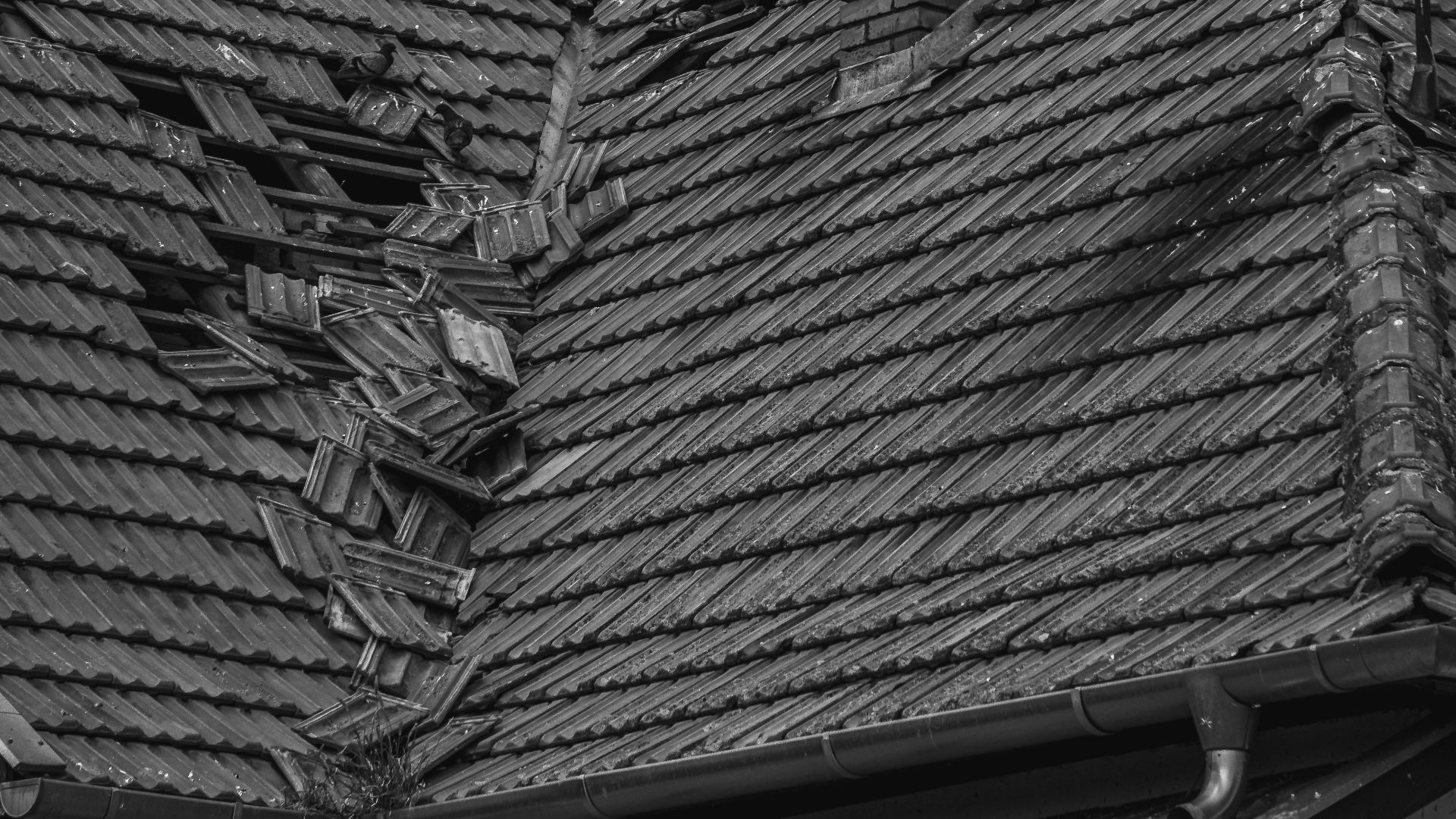 A grayscale photo of a broken roof with a chimney and antenna under a cloudy sky.