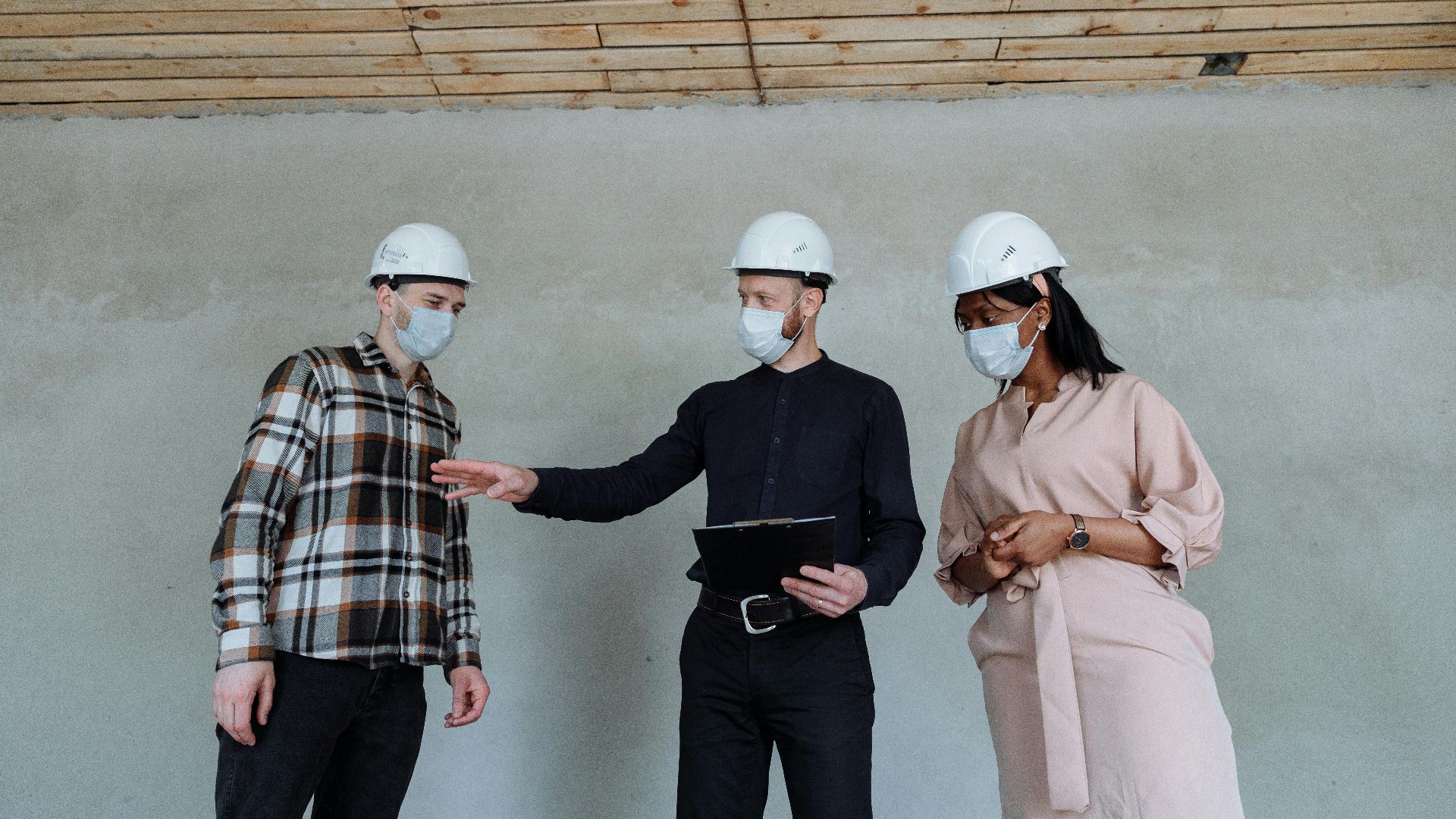 Three engineers wearing hard hats and masks discuss a project inside a construction site.