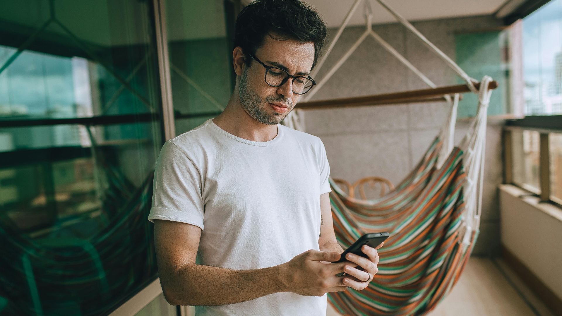 Young man with glasses using a smartphone on a balcony with a hammock.