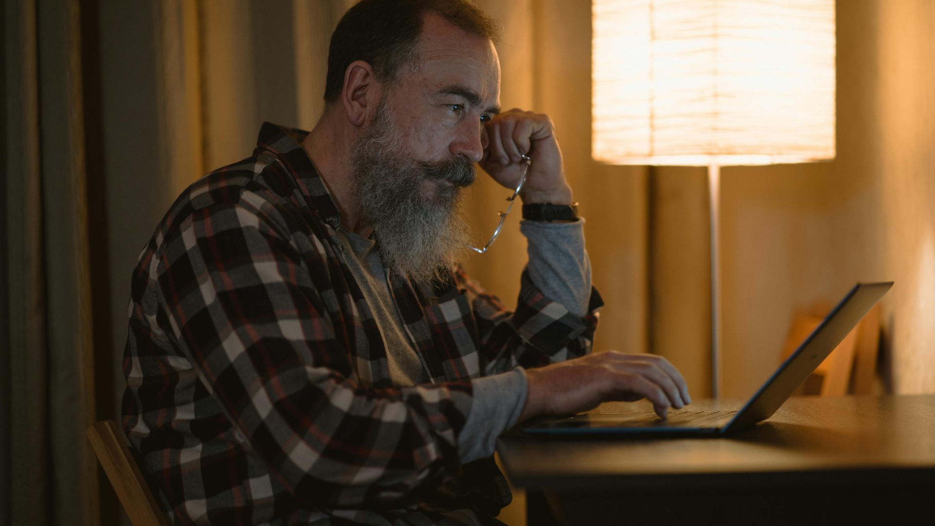 Elderly man with a beard working late on laptop under warm lamp light indoors.