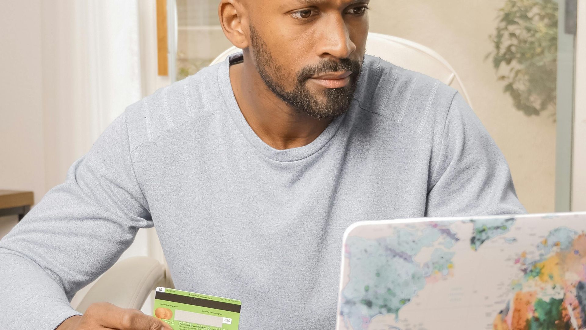 Bald man using credit card for online shopping on colorful world map laptop indoors.