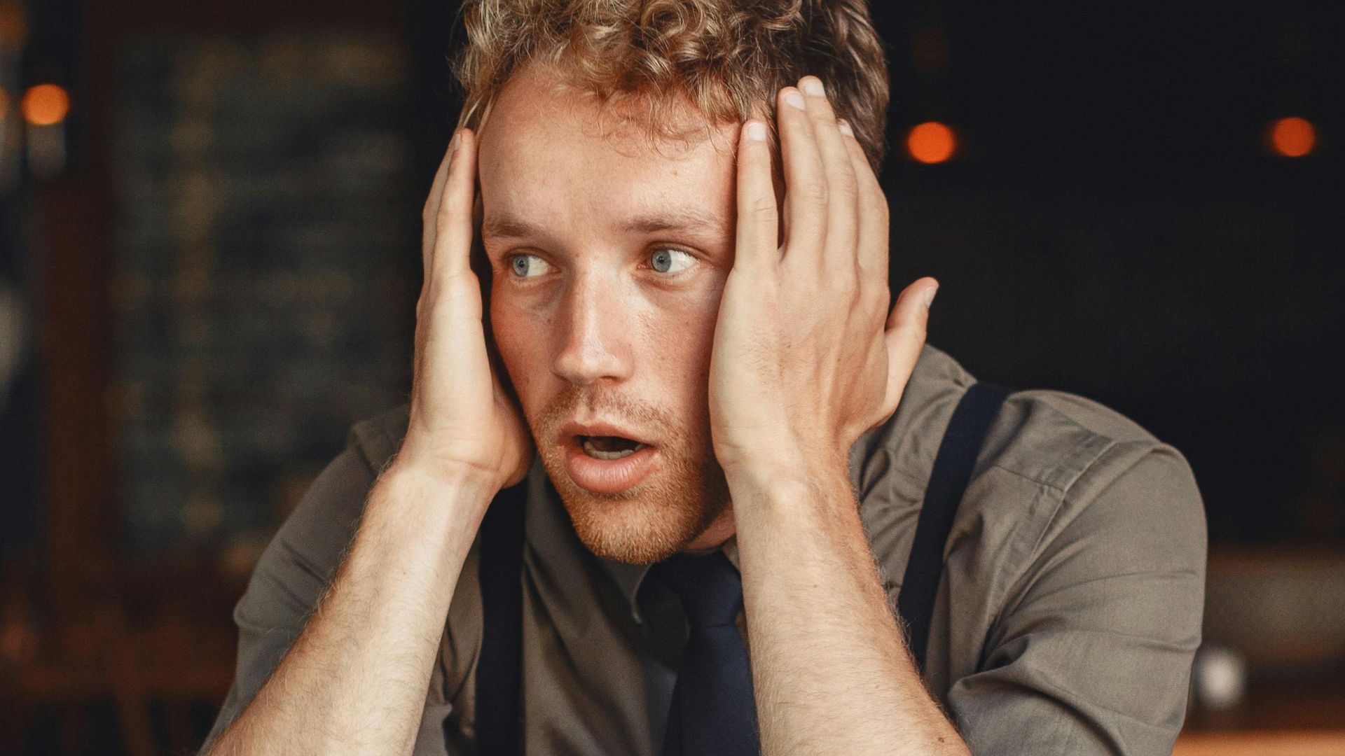 A stressed man with hands on head, surrounded by work materials, sits in a dimly lit restaurant.