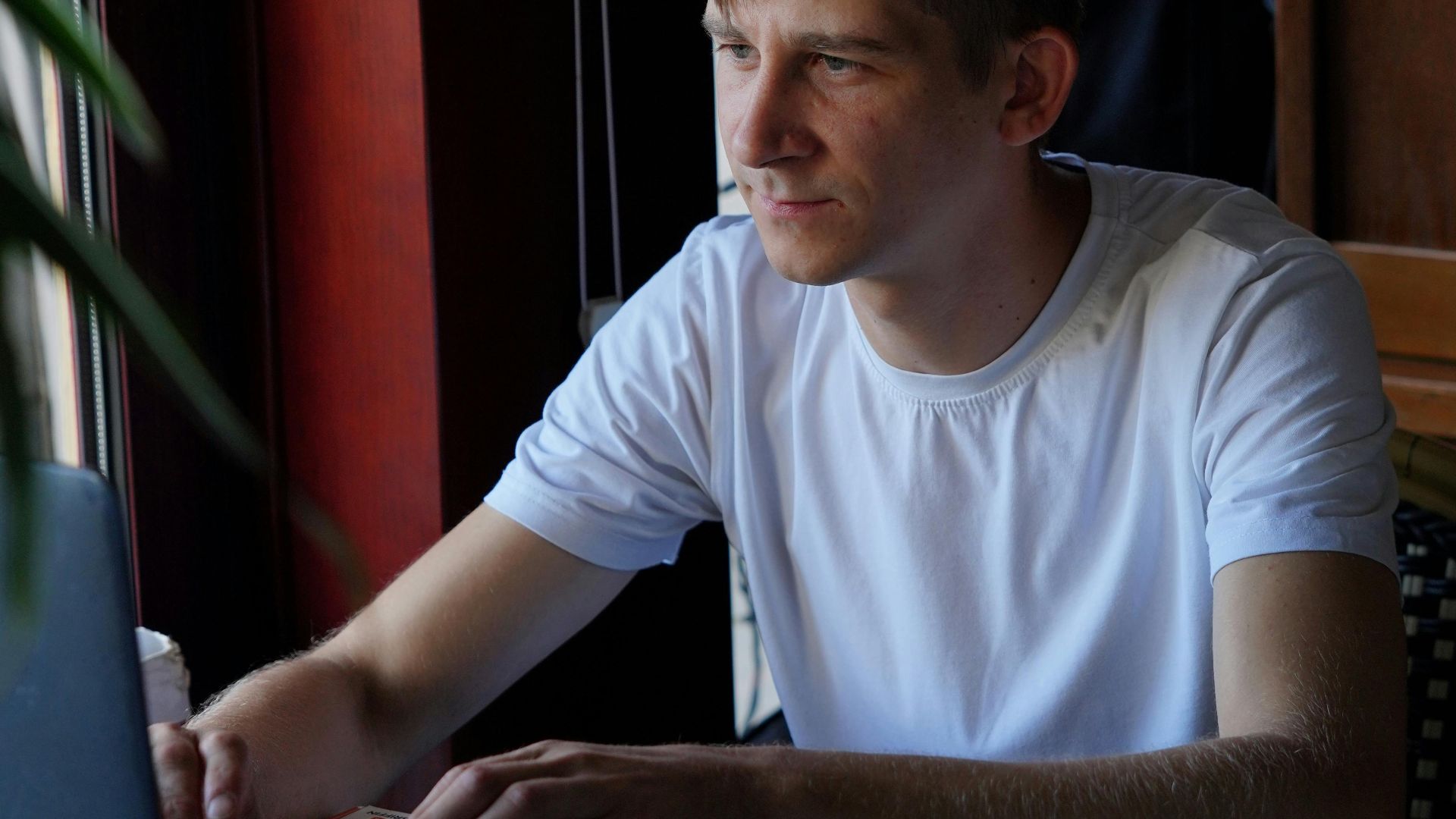 A young man in a café working on his laptop by the window, focused and casual.