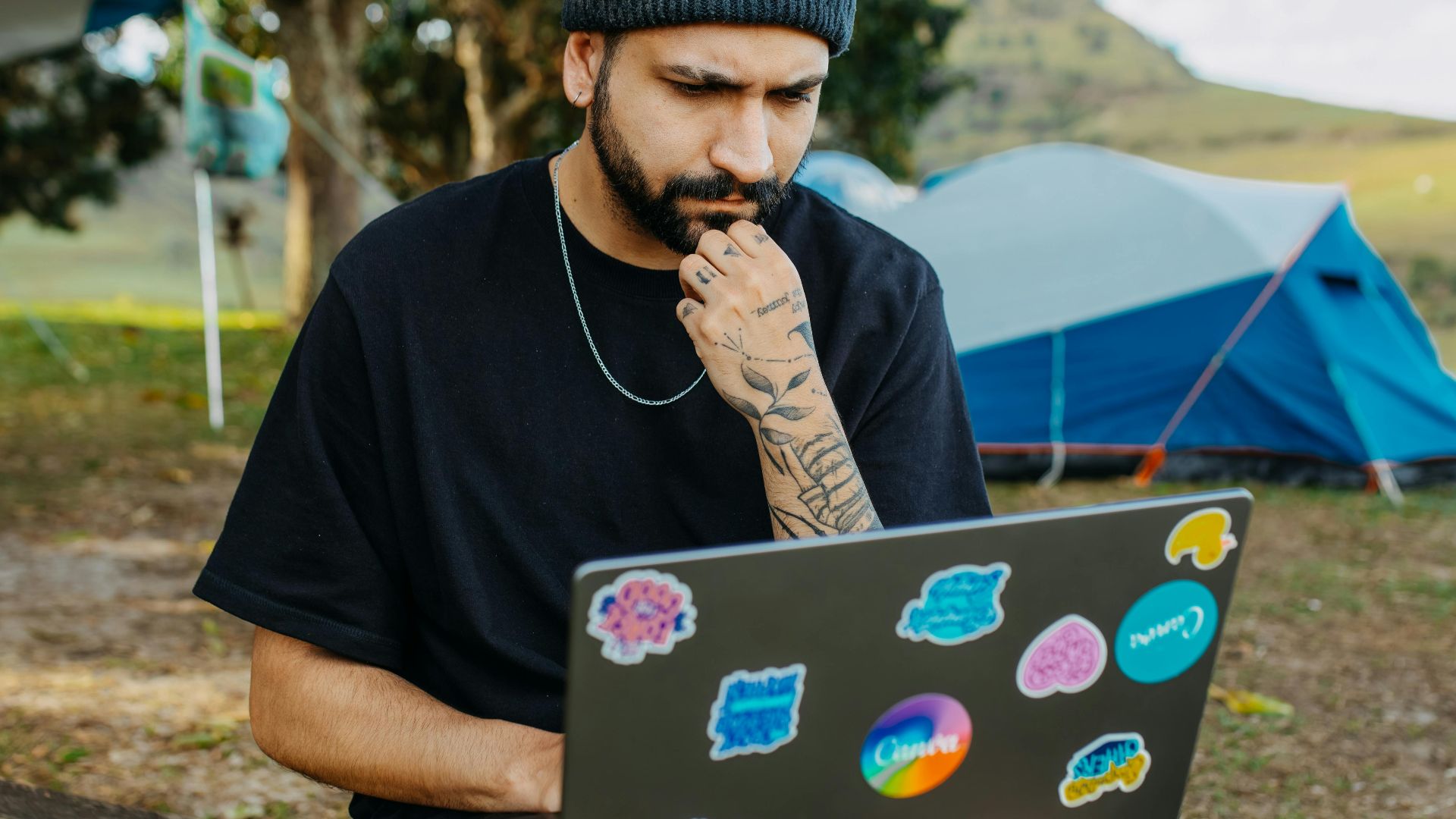 A man with a beard sits outdoors, working on a laptop at a campsite, surrounded by nature.