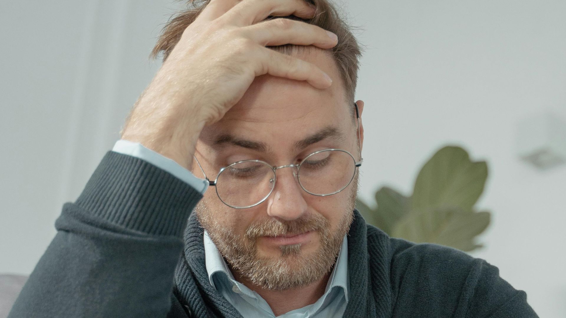 A thoughtful man with glasses sits indoors, hand on head, reflecting stress or contemplation.
