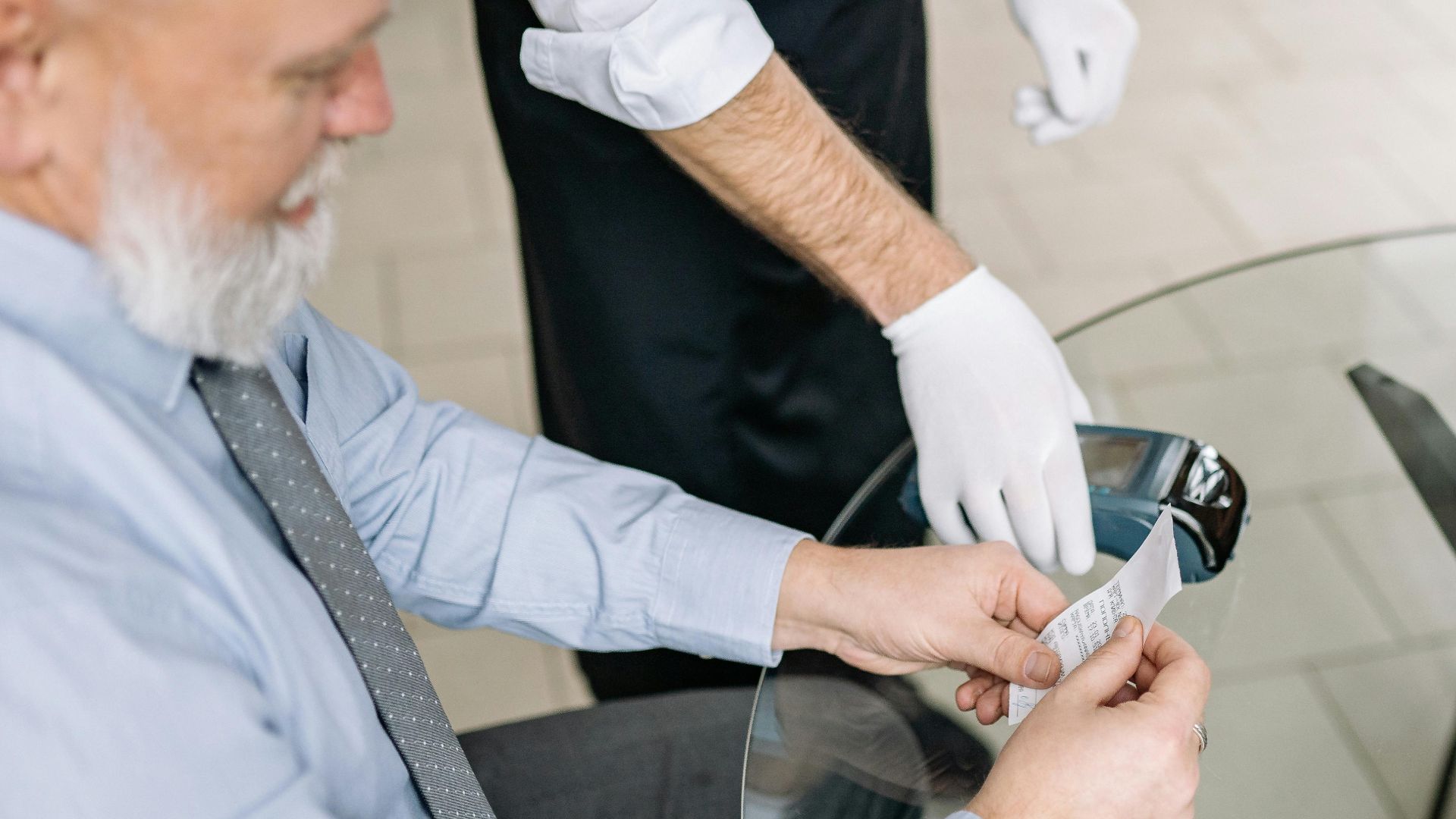 Senior man making a payment via credit card with a waiter in gloves assisting.