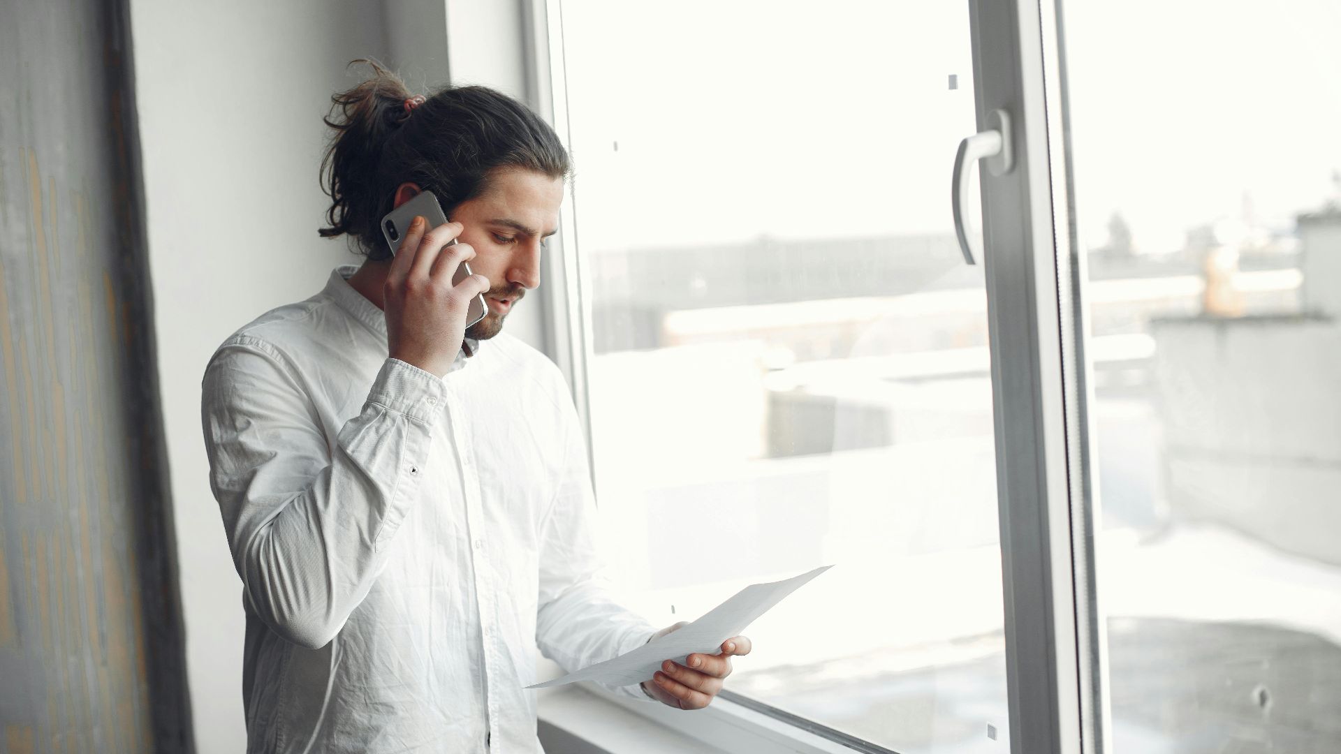 Young man in white shirt, on phone call holding a document, standing by a large window.