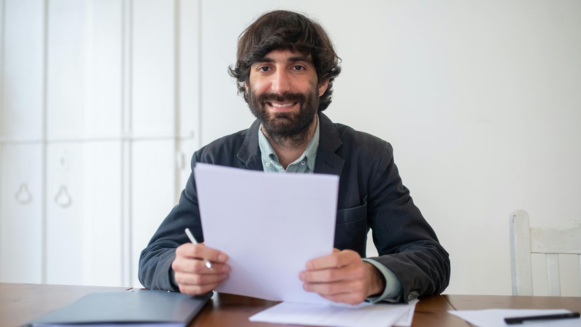Smiling businessman in a black jacket reviewing documents at his desk, indoors.
