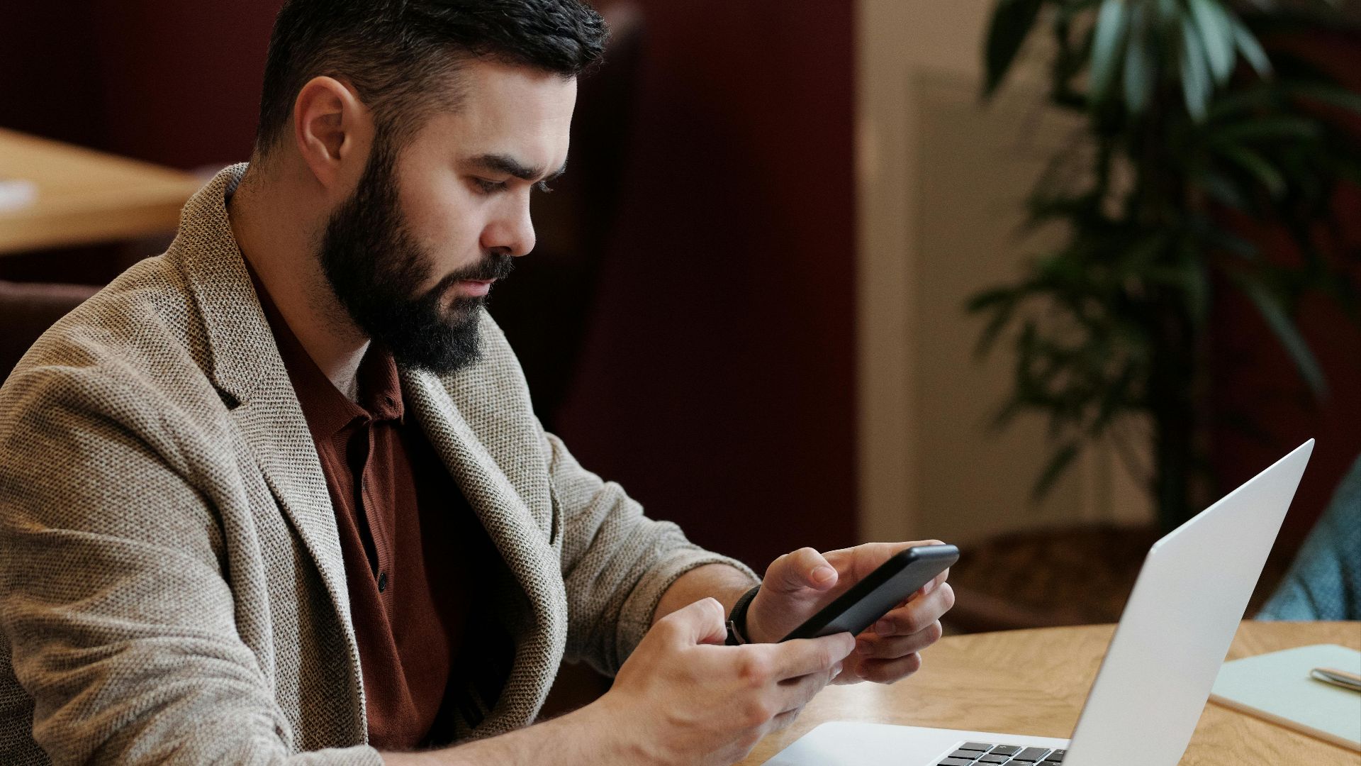 Bearded man using smartphone and laptop at café with coffee, focused on remote work.