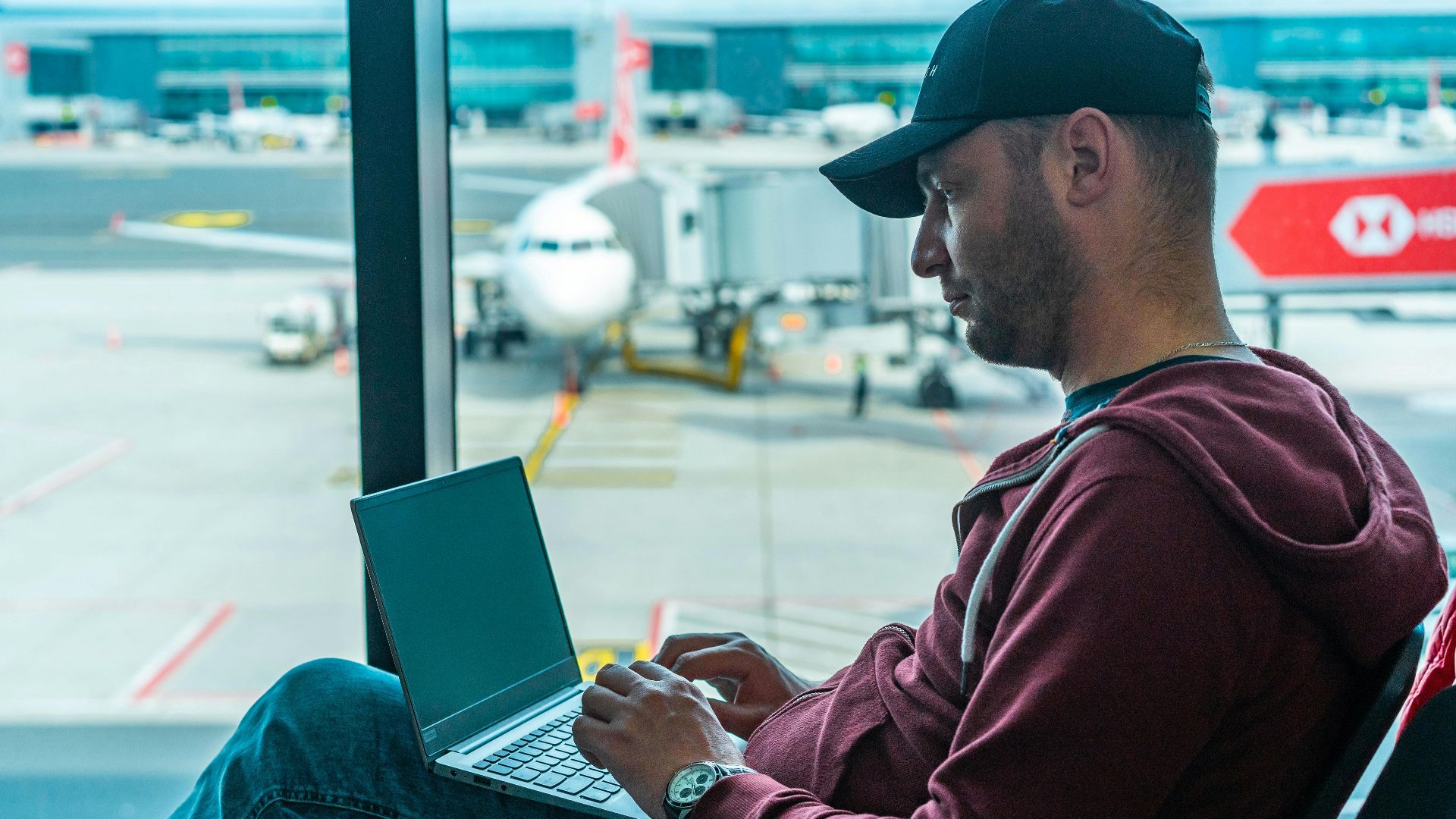 A man working on his laptop in an airport terminal, with a plane visible outside.