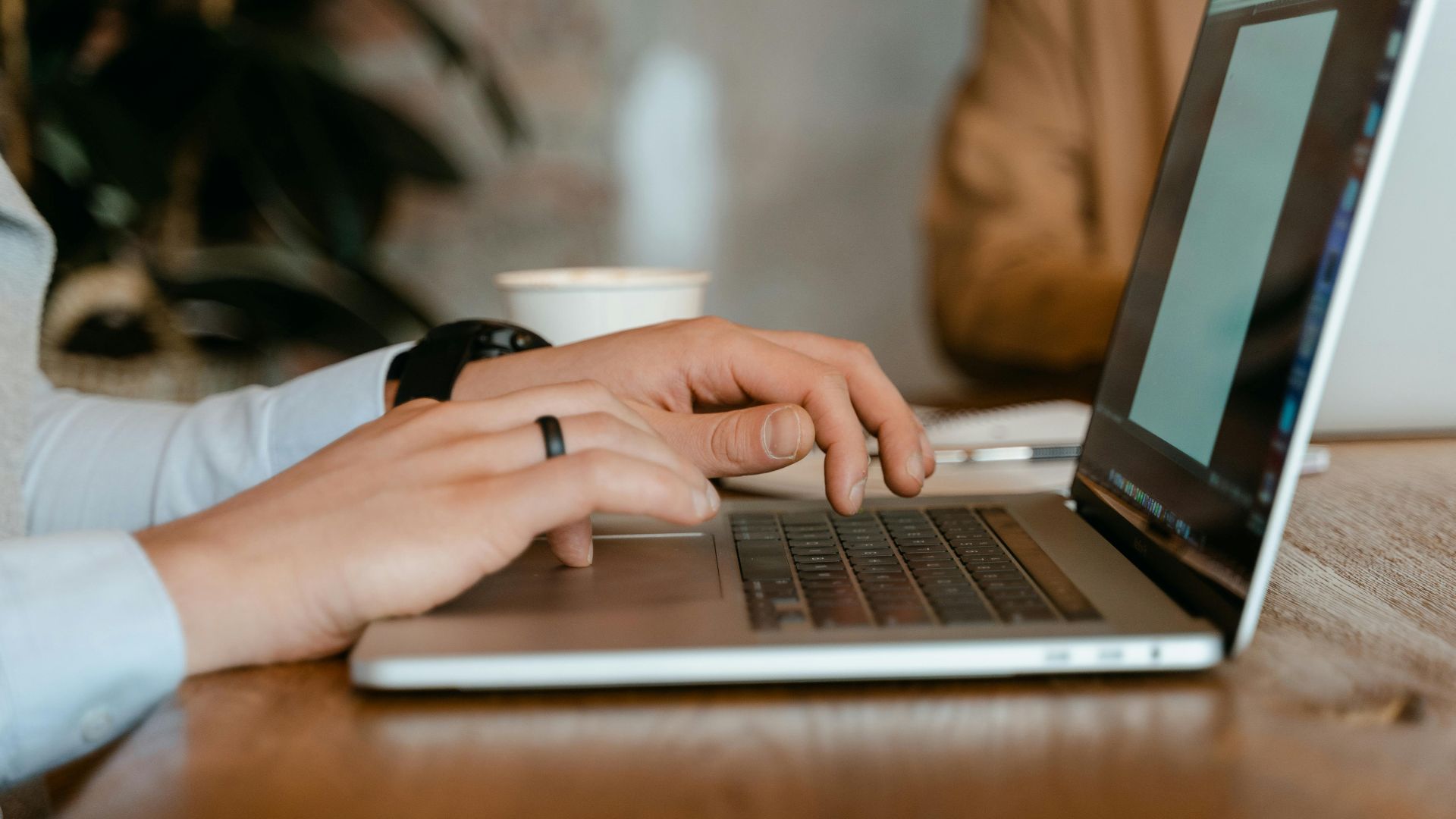 A person typing on a laptop at a wooden table in a modern office setting.