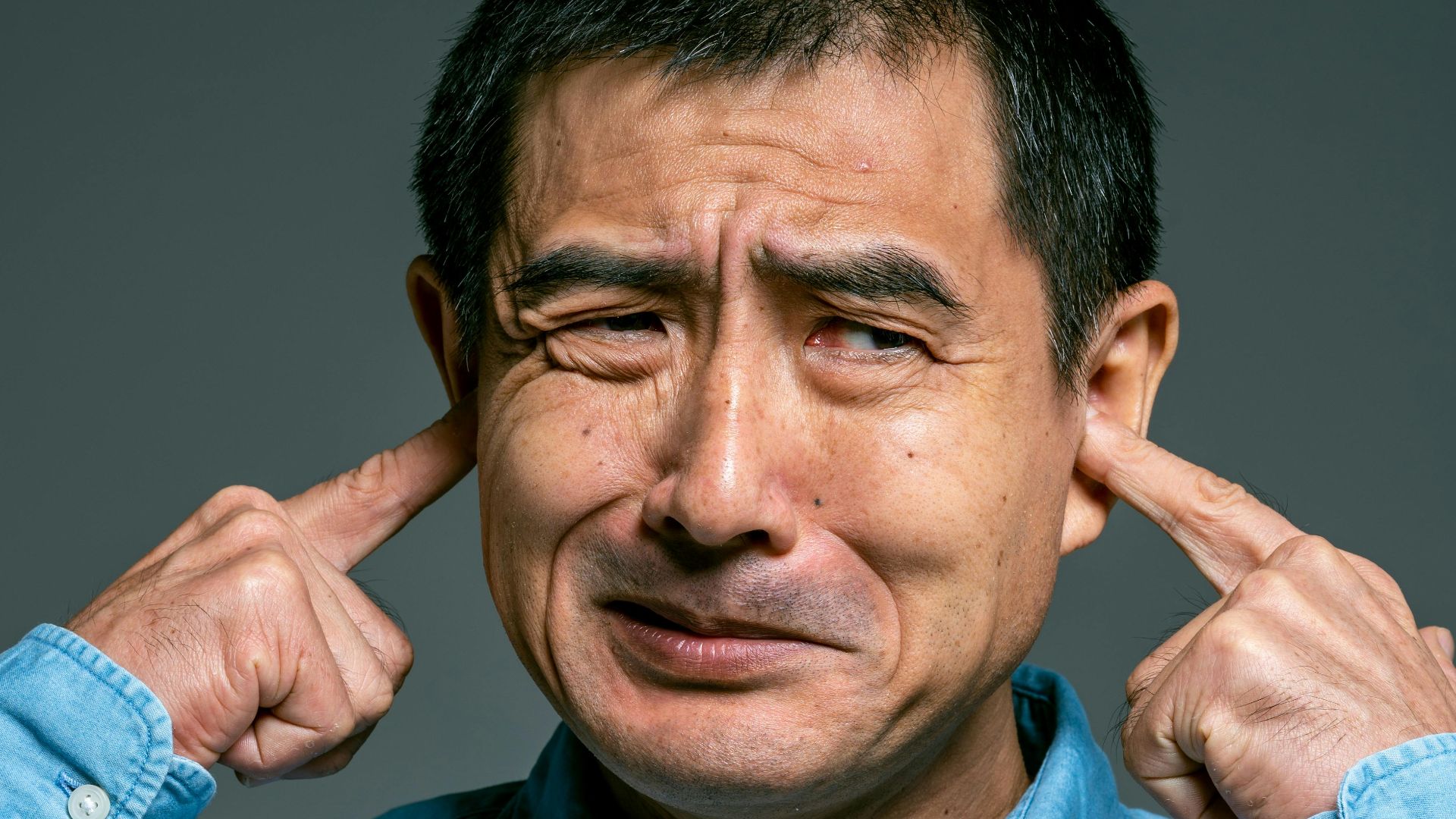 Asian man in blue denim shirt expressing frustration by covering his ears in a close-up portrait.