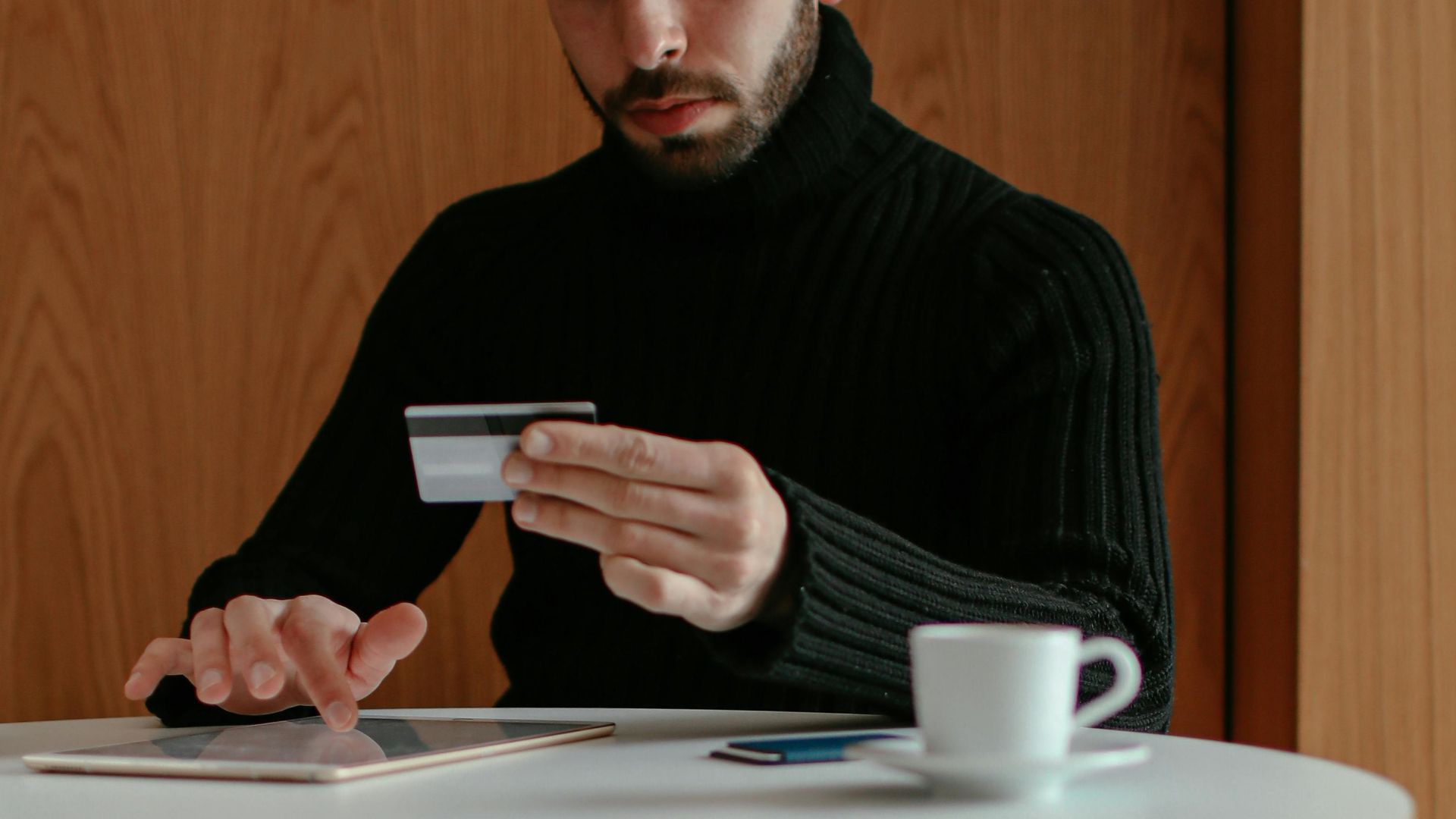Concentrated young bearded male sitting at table with cup of coffee and browsing tablet while doing online purchase with credit card