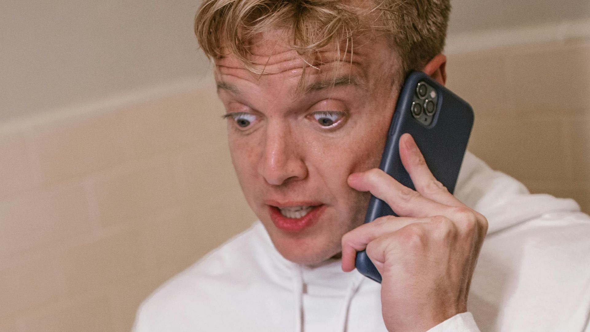 Young man talking on phone while using laptop in bathroom setting.