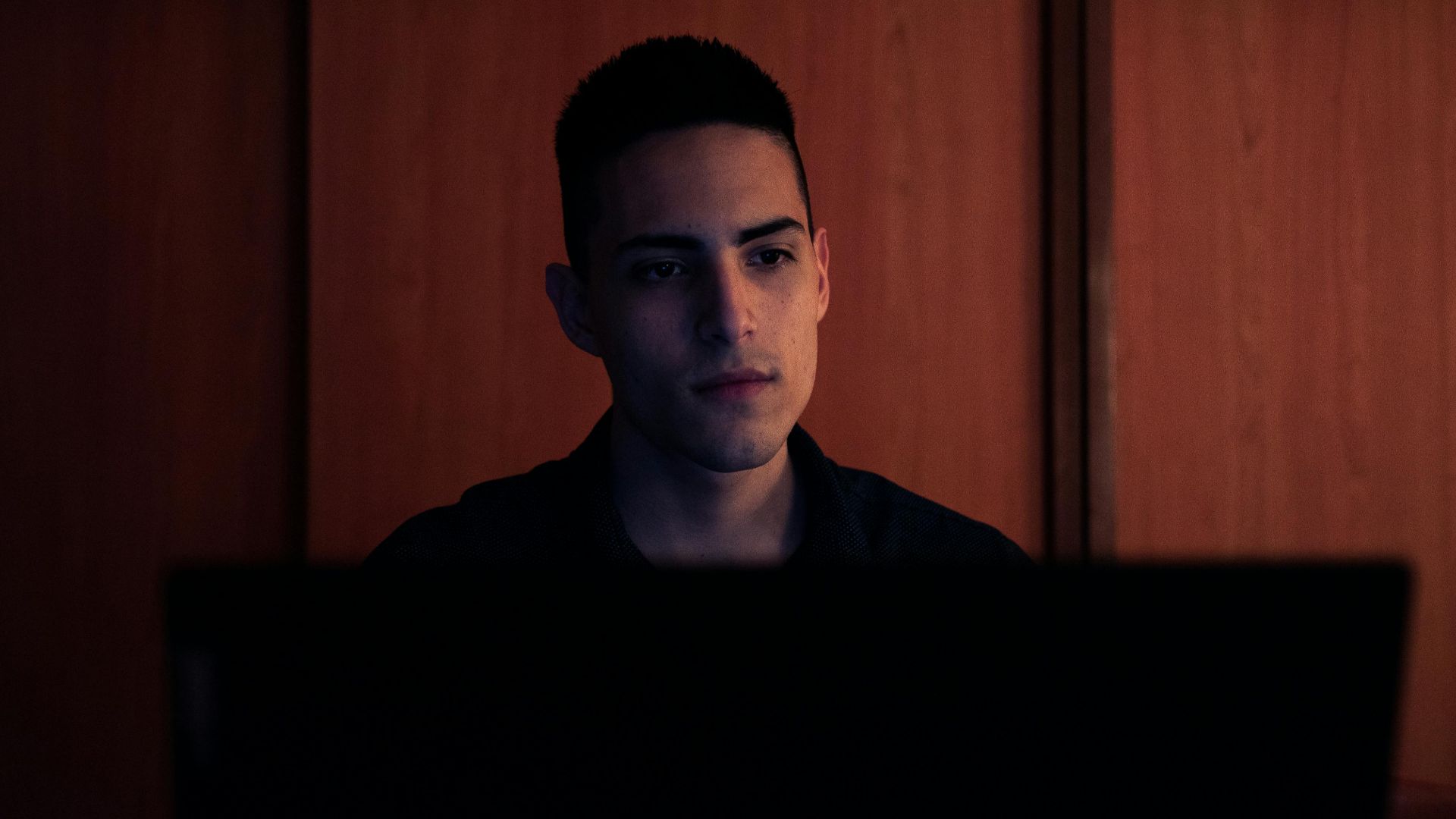 A young man working on his laptop, focused and serious, in an indoor setting with warm lighting.