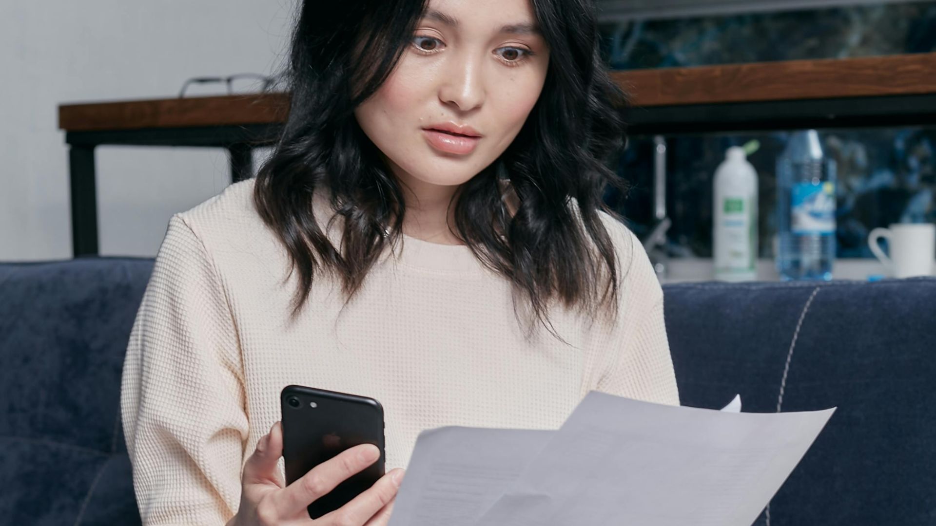 Young woman reviews documents and phone indoors, showcasing concern while seated on a couch with scattered papers. Business concept.