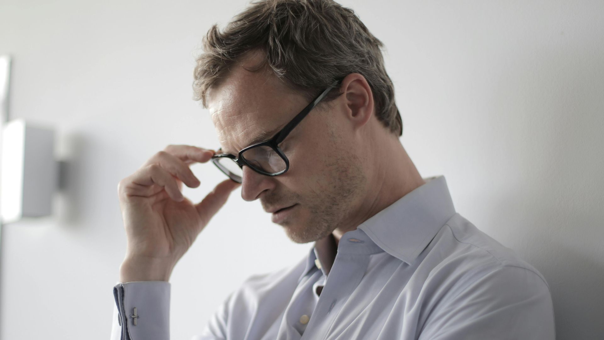 An adult man in a shirt and glasses appears stressed while leaning against a wall indoors.