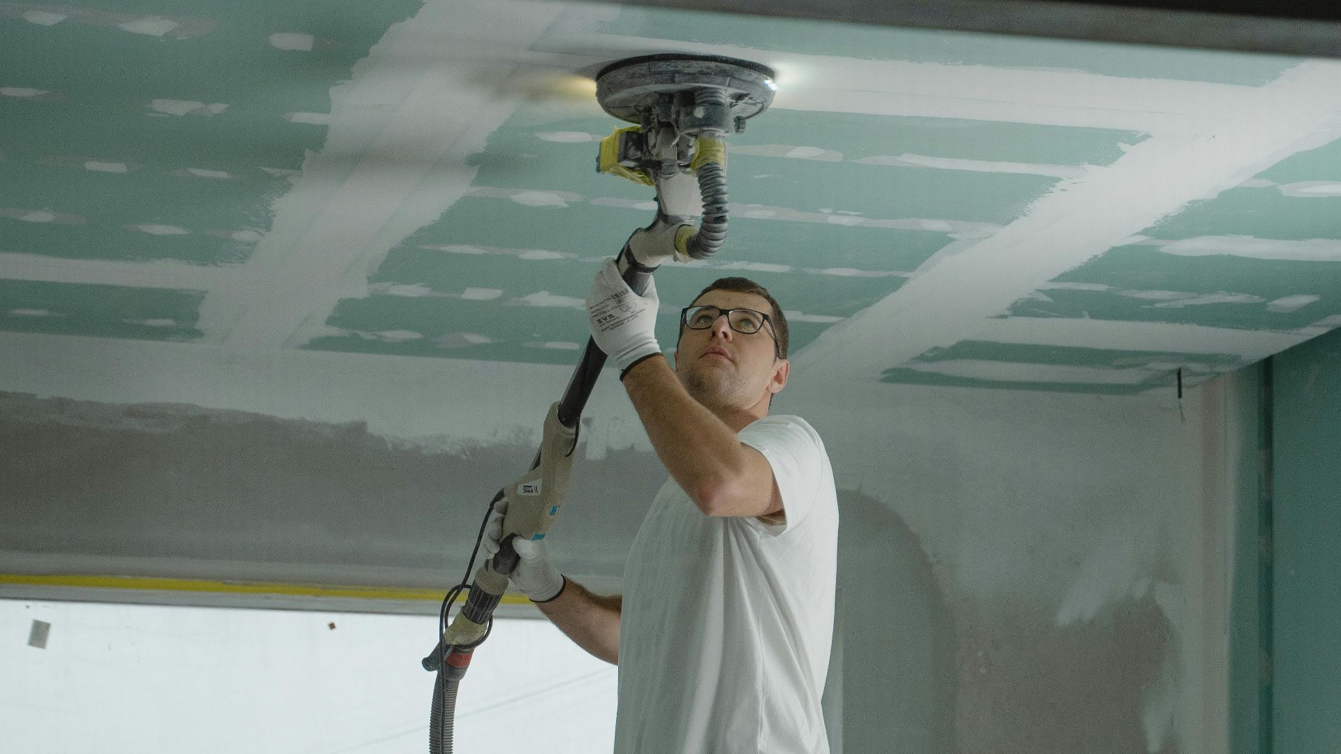 A man on stilts uses a ceiling sander for home renovation, focusing on drywall preparation.