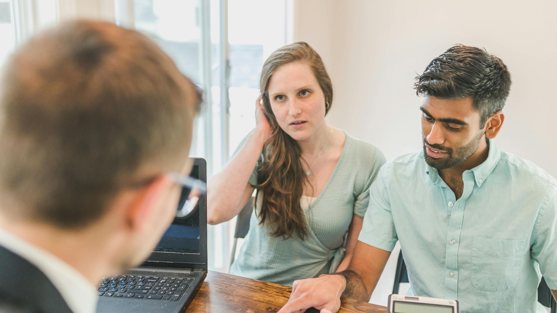 A young couple consults with a realtor about buying a home, indoors in a modern office setting.
