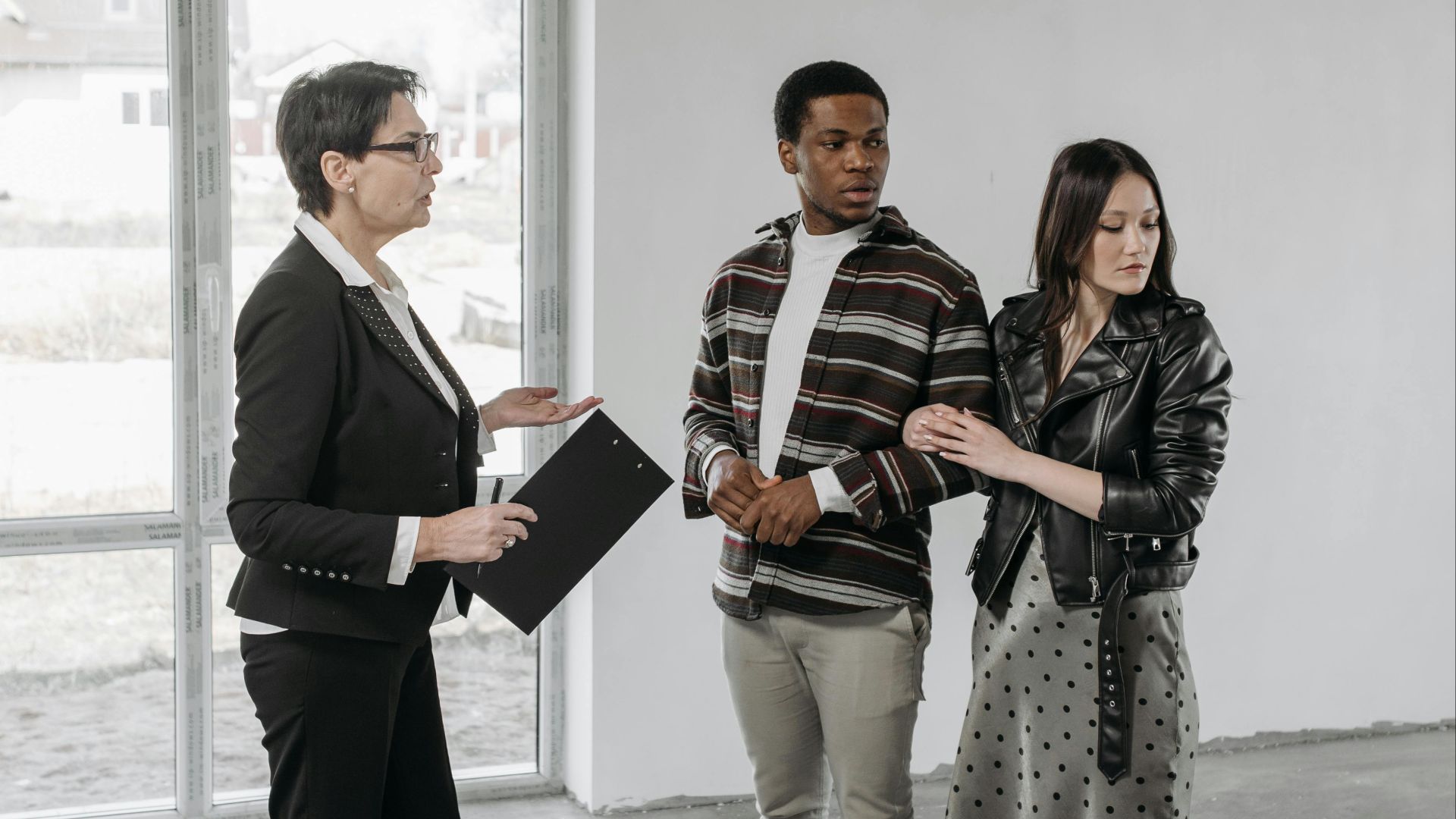 Multicultural couple discussing potential house purchase with realtor inside an empty room.
