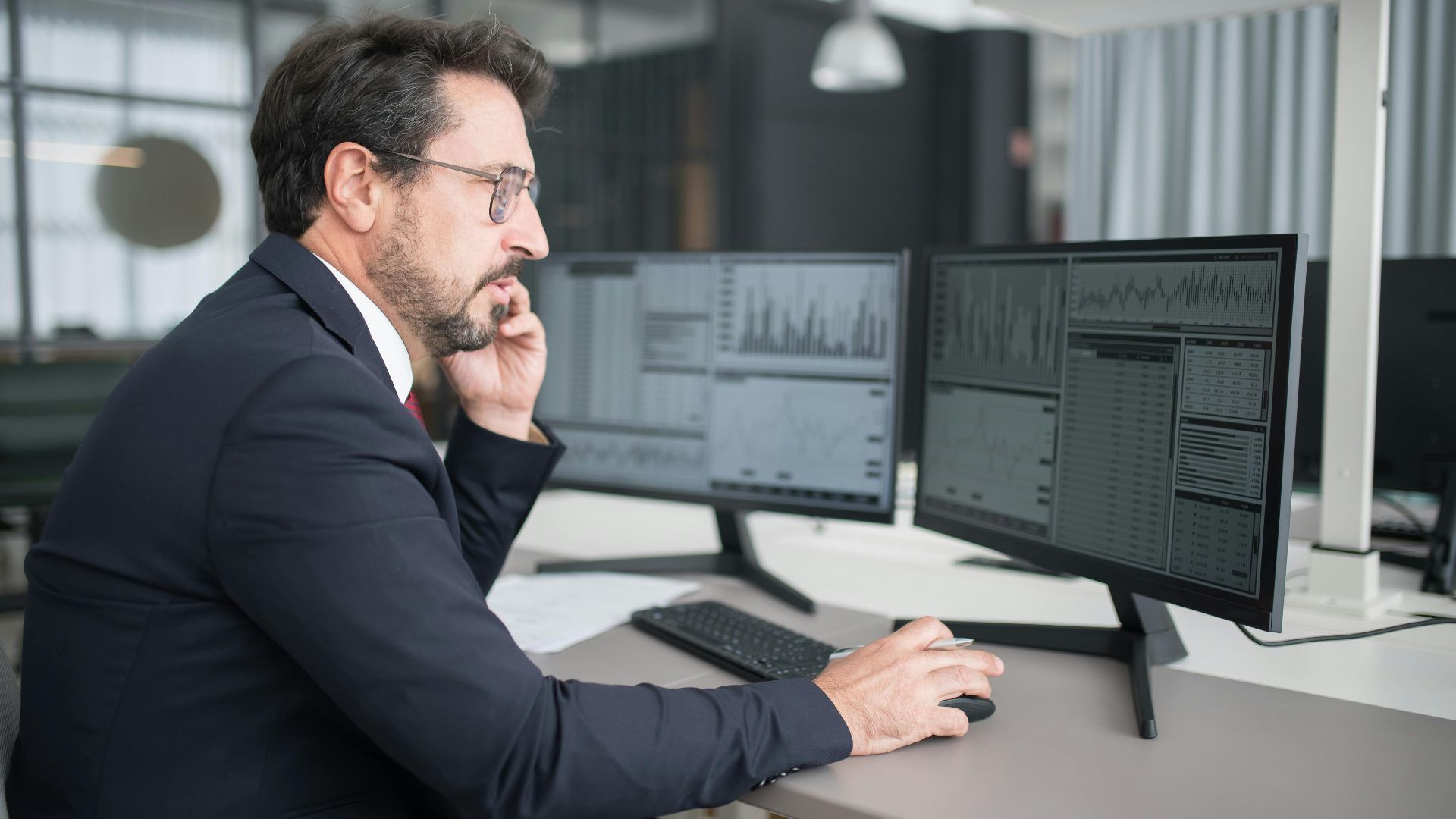 Businessman analyzing stock market data on dual monitors in a modern office setting.