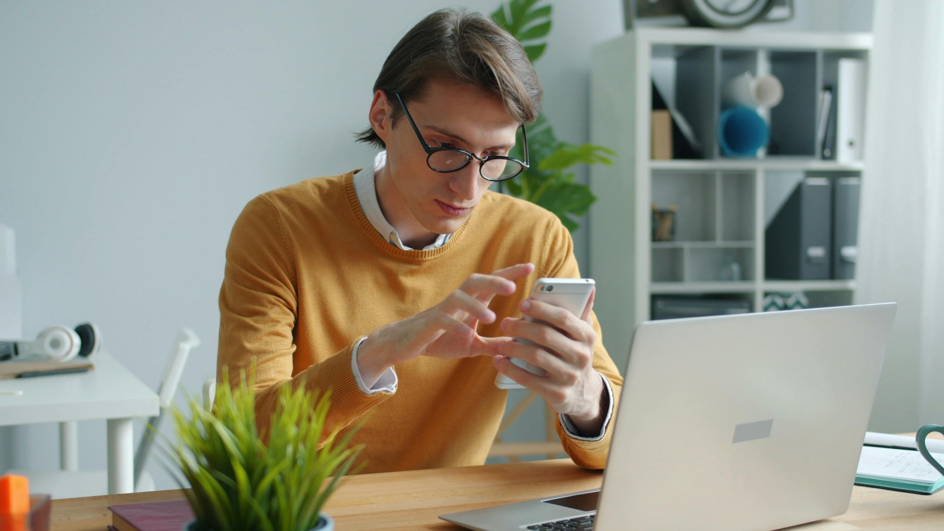 A young adult working on a smartphone and laptop in a modern office.