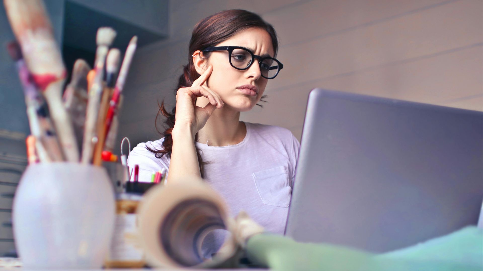 Focused woman with glasses working on laptop in an artistic workspace with paintbrushes.
