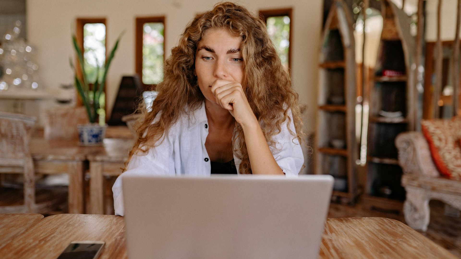 A thoughtful woman working remotely on a laptop in a cozy indoor setting.