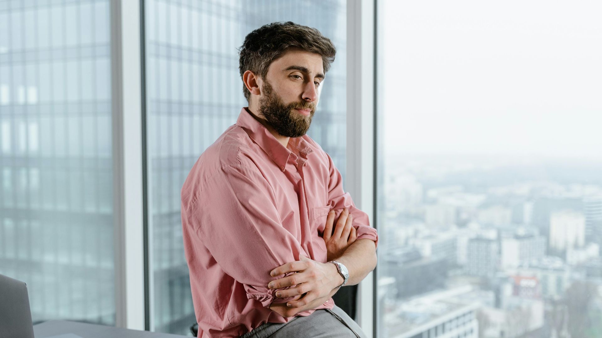 Man in pink shirt sitting on table, arms crossed, overlooking city from high-rise office.