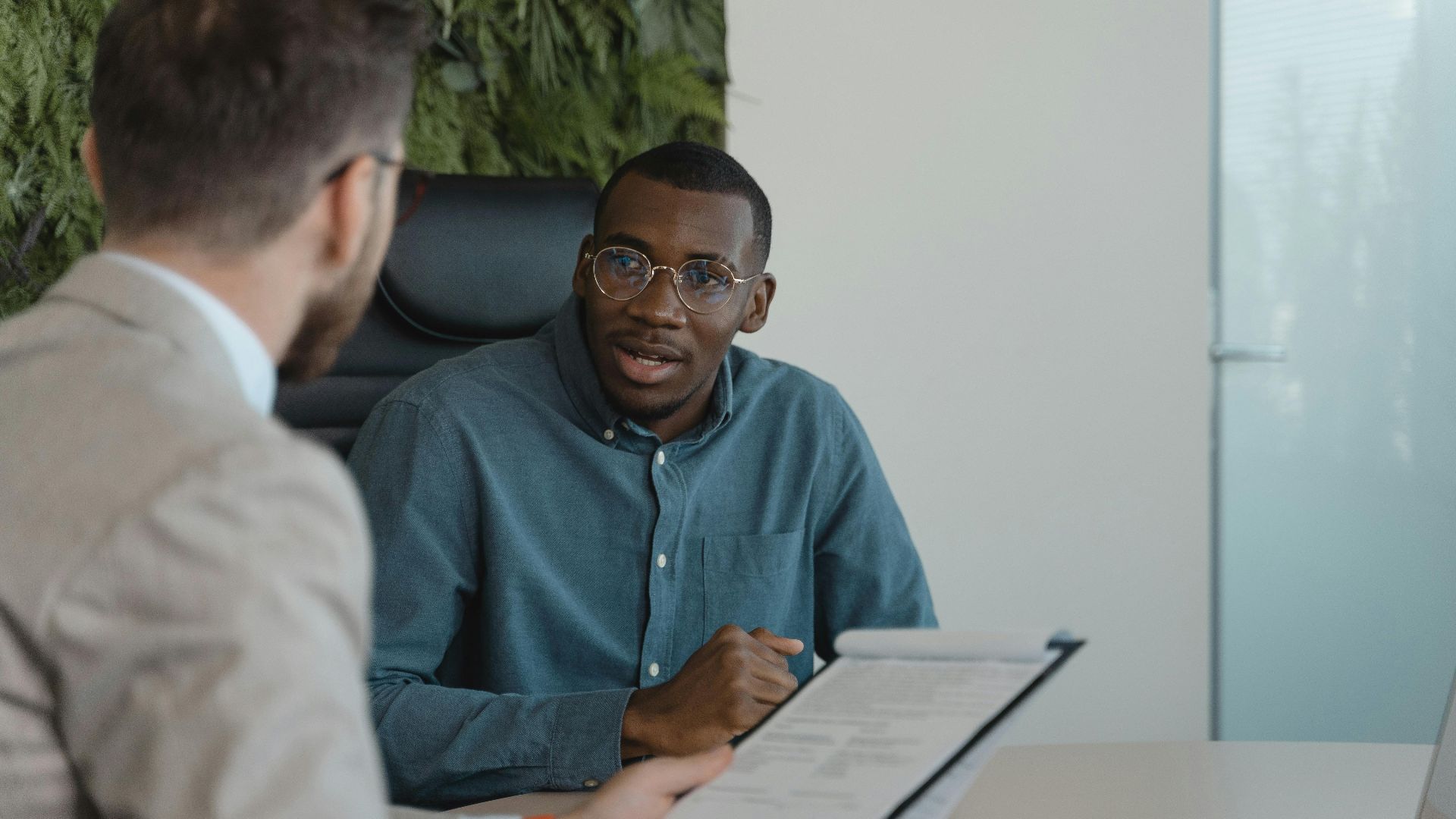 Two men engaged in a professional meeting at an office table with documents and a plant wall background.