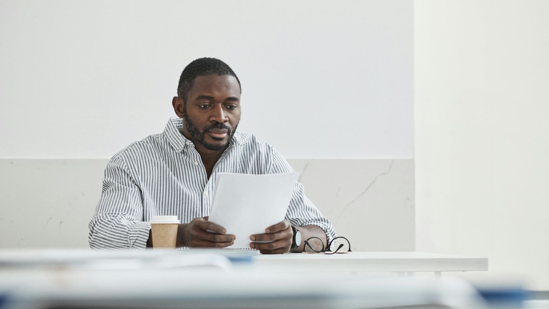 African American man sitting indoors, reading papers with a coffee cup nearby.