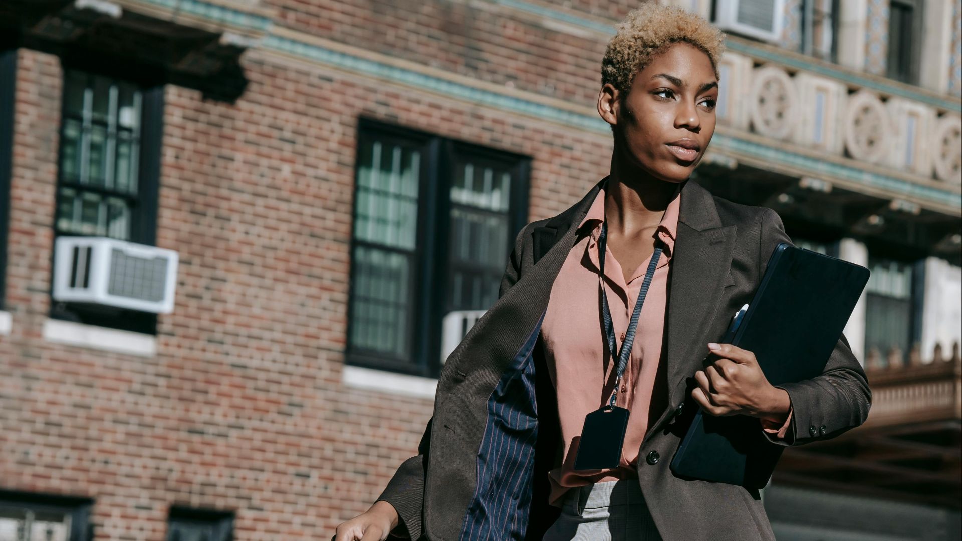 Self assured young African American female entrepreneur with short dyed hair in stylish formal outfit walking on city street with laptop in hand and looking away