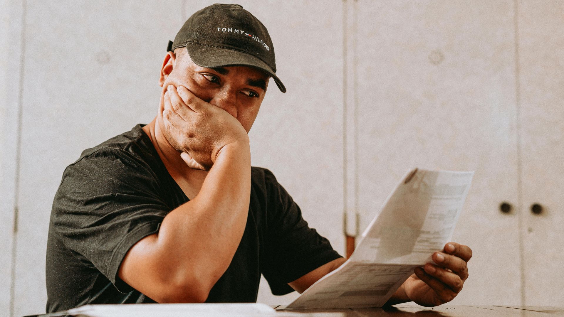 A man sits indoors, deeply focused on reviewing paperwork, expressing concern and contemplation.