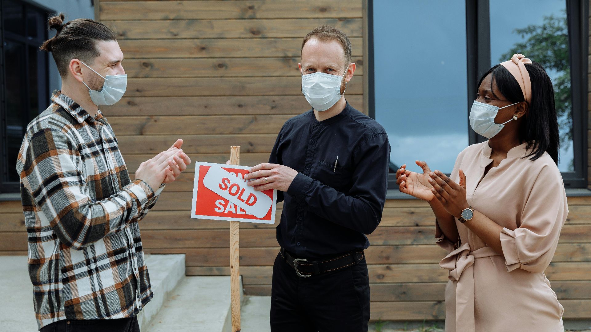 Real estate agent and clients celebrate sale with masks and a sold sign outdoors.