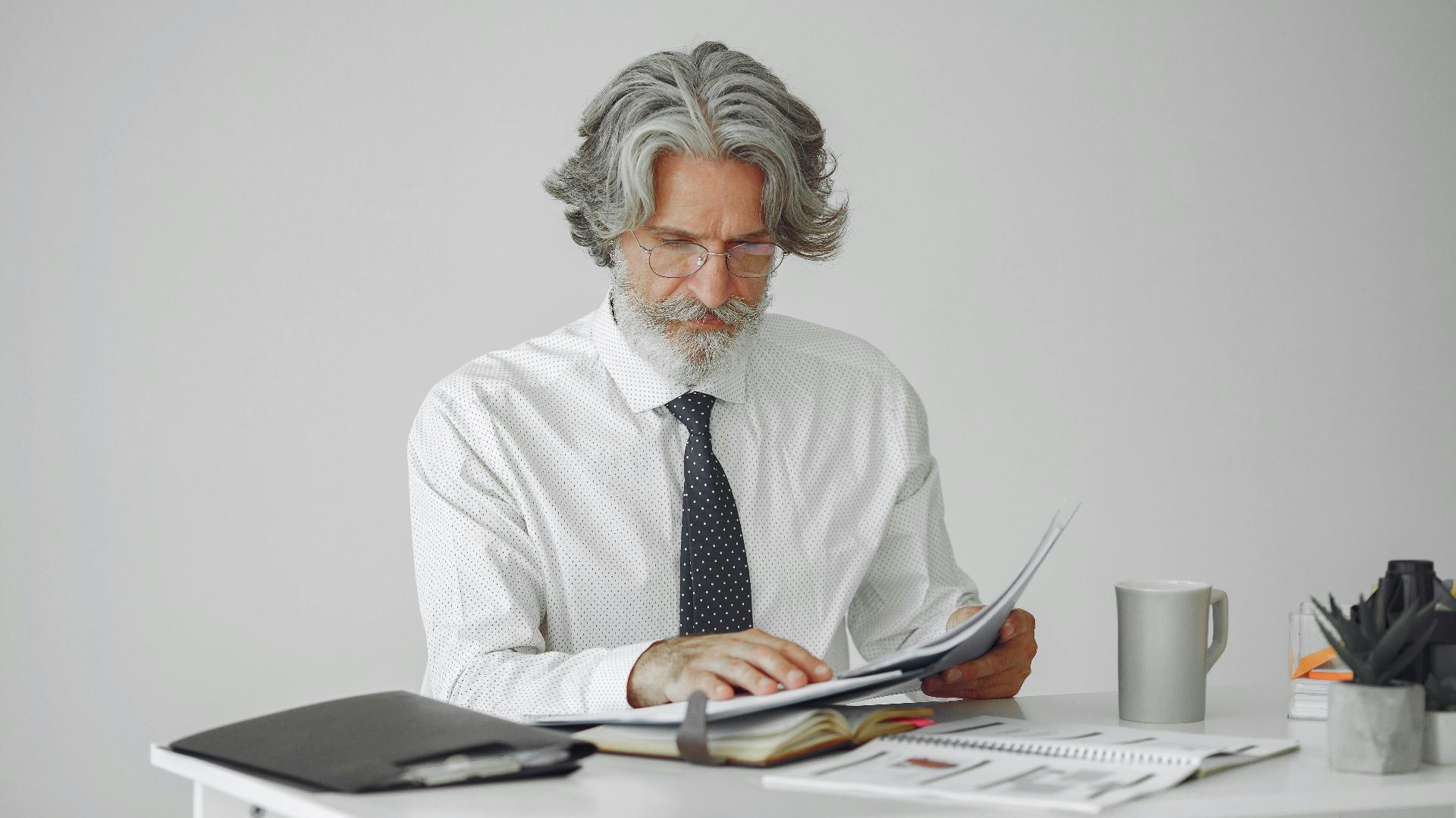 An elderly businessman reviewing documents at his desk in a modern office setting.