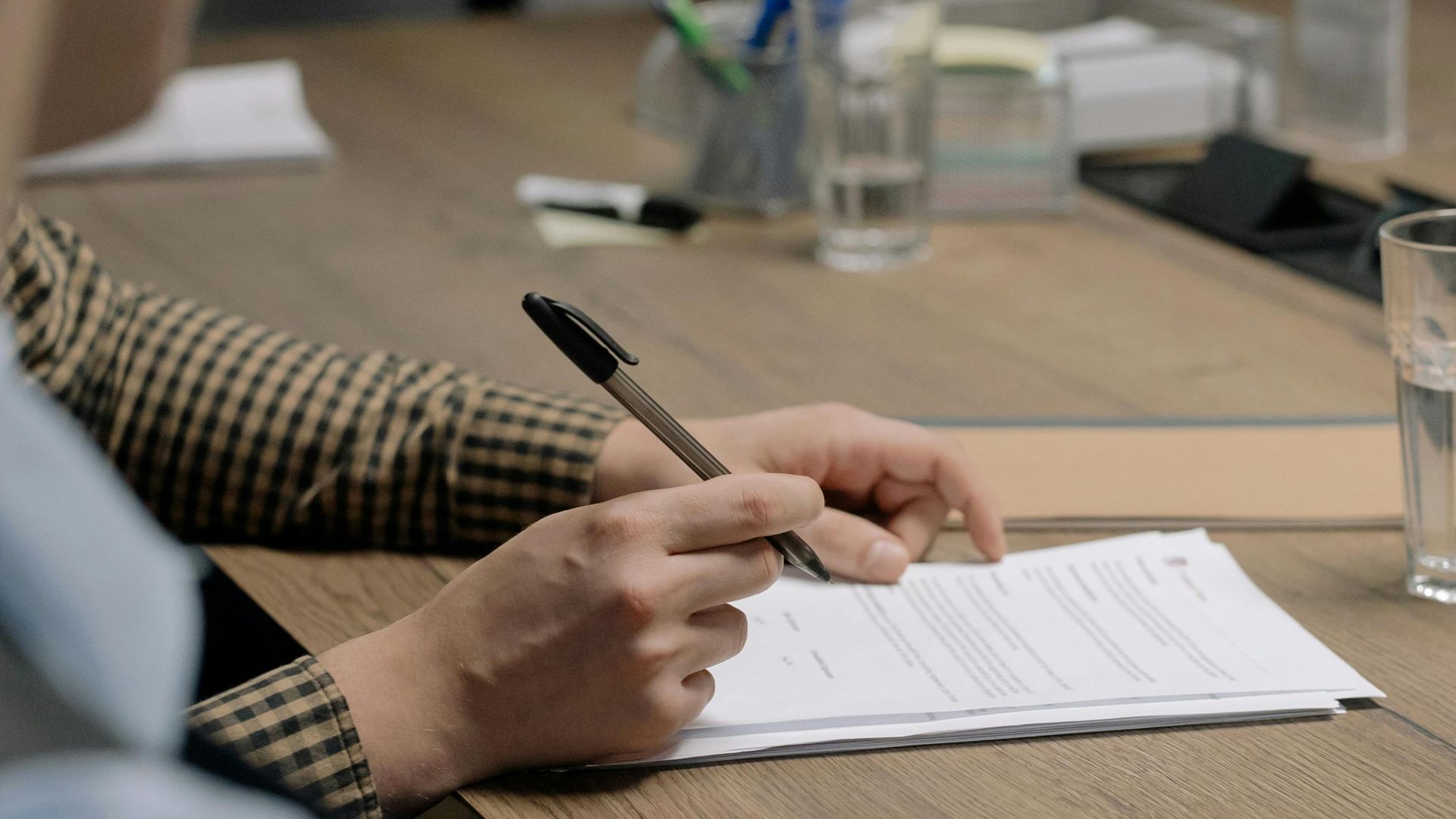 Person in business attire signing a document at a wooden table in an office setting.