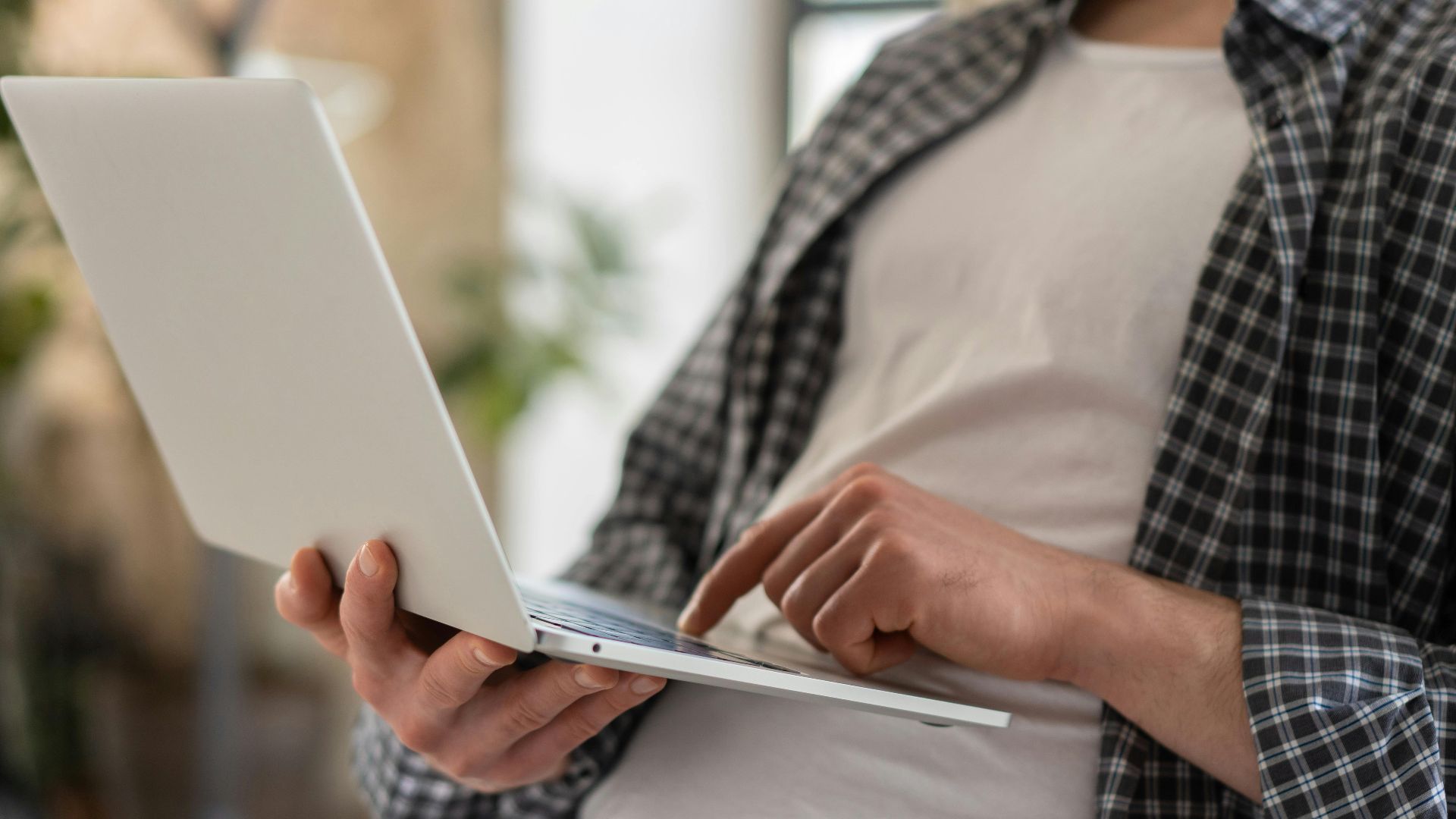 Young adult in casual shirt using a laptop indoors with blurred background and shallow focus.
