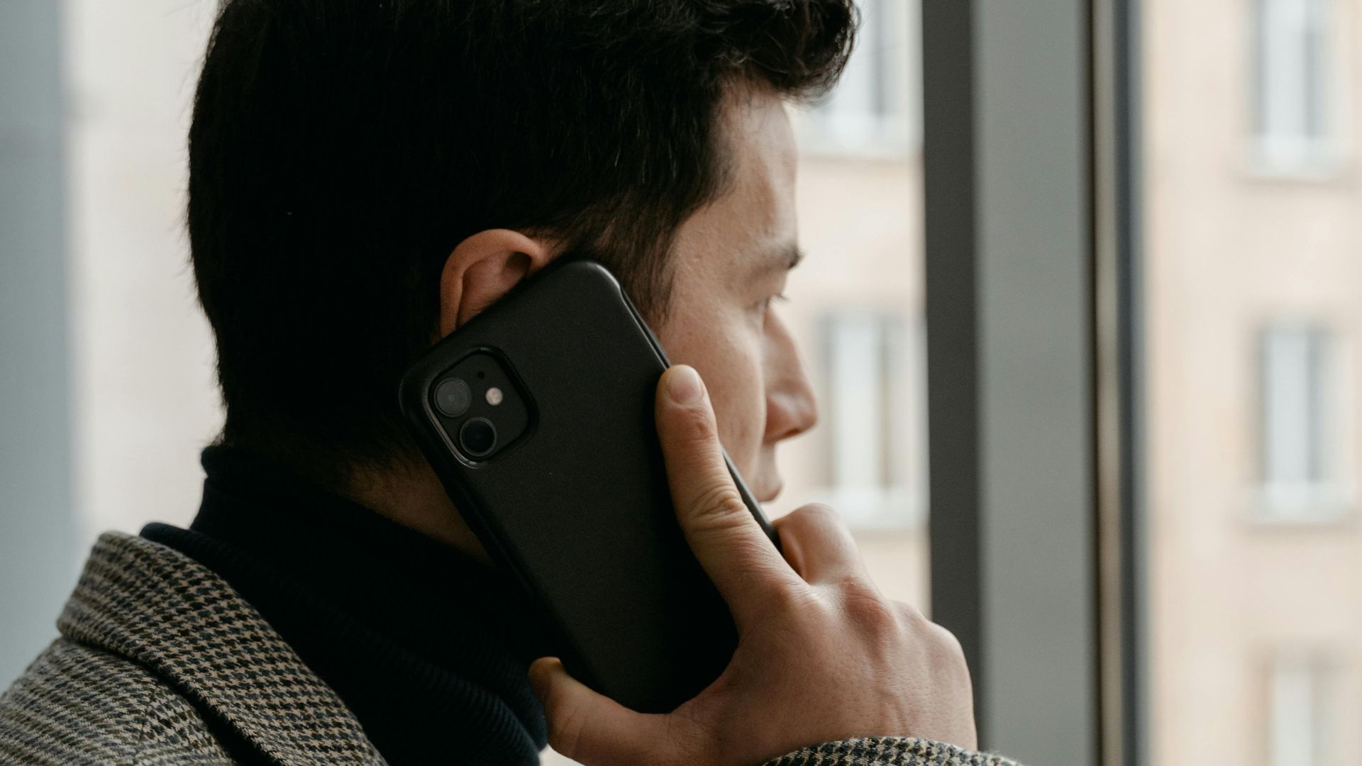 Man in a checkered blazer using a smartphone while gazing out a window.