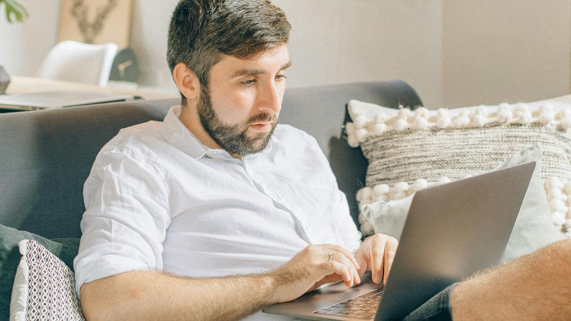 Adult man sitting on couch using laptop, working remotely from cozy home setting.