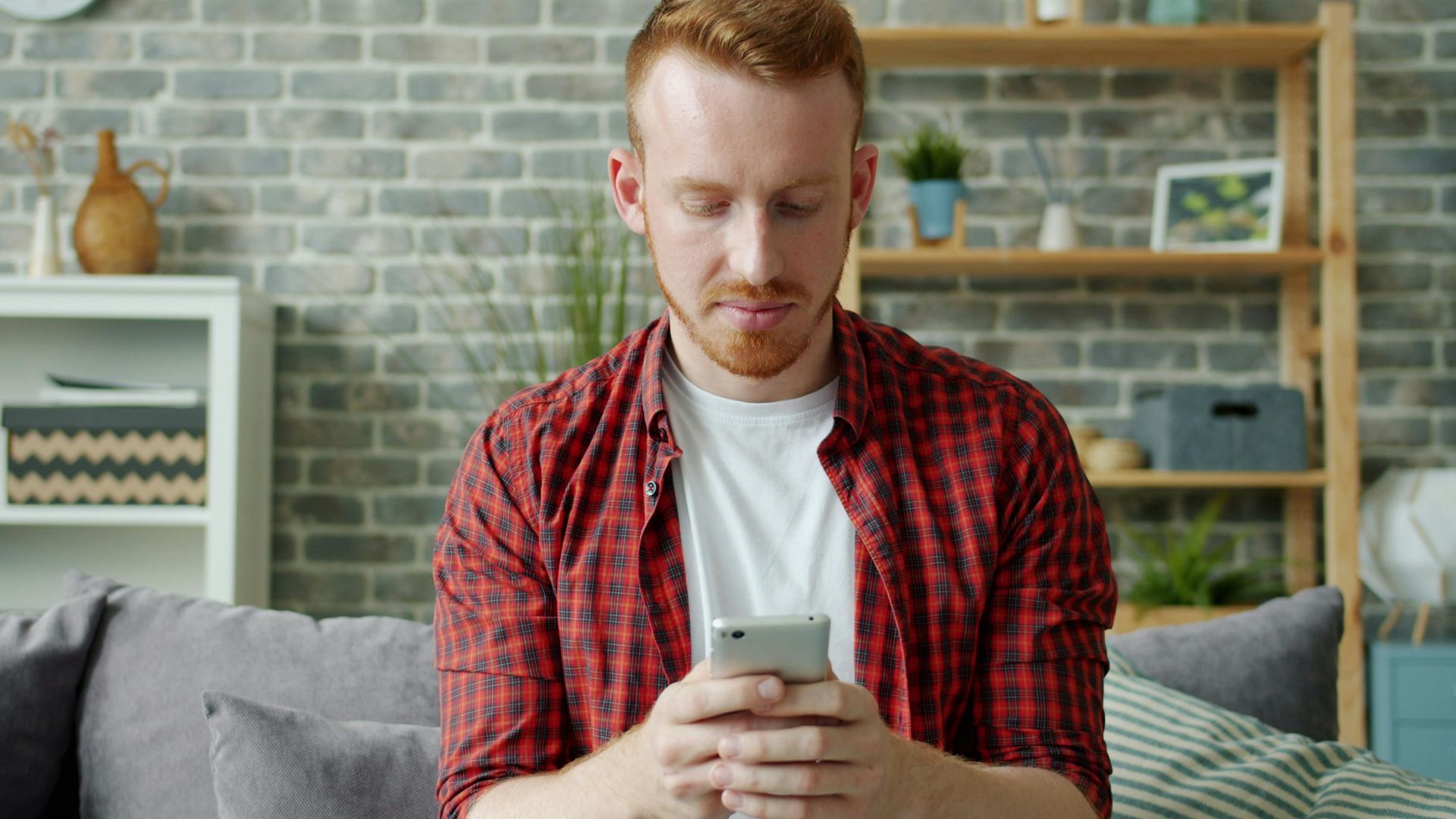 Man in red plaid shirt texting on smartphone at home, modern lifestyle.