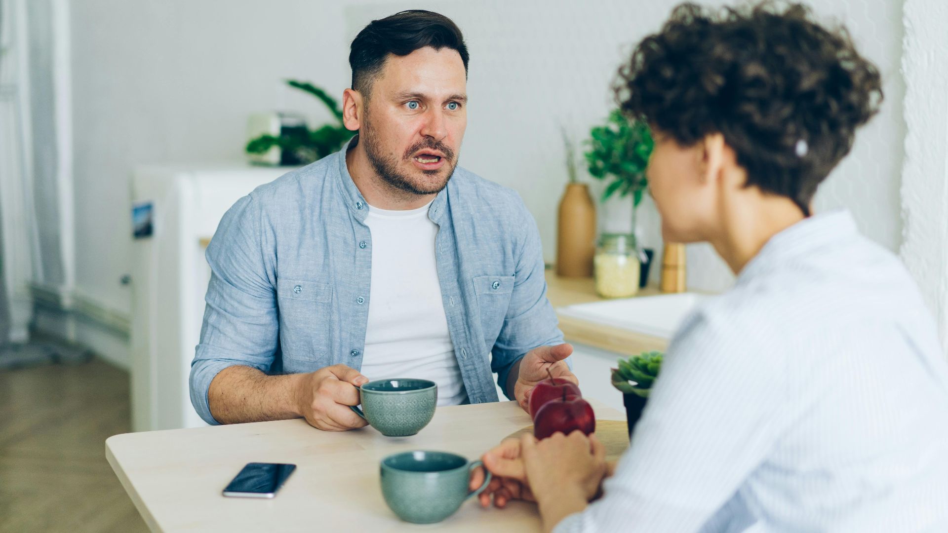 Couple having a serious conversation at the kitchen table with coffee cups.